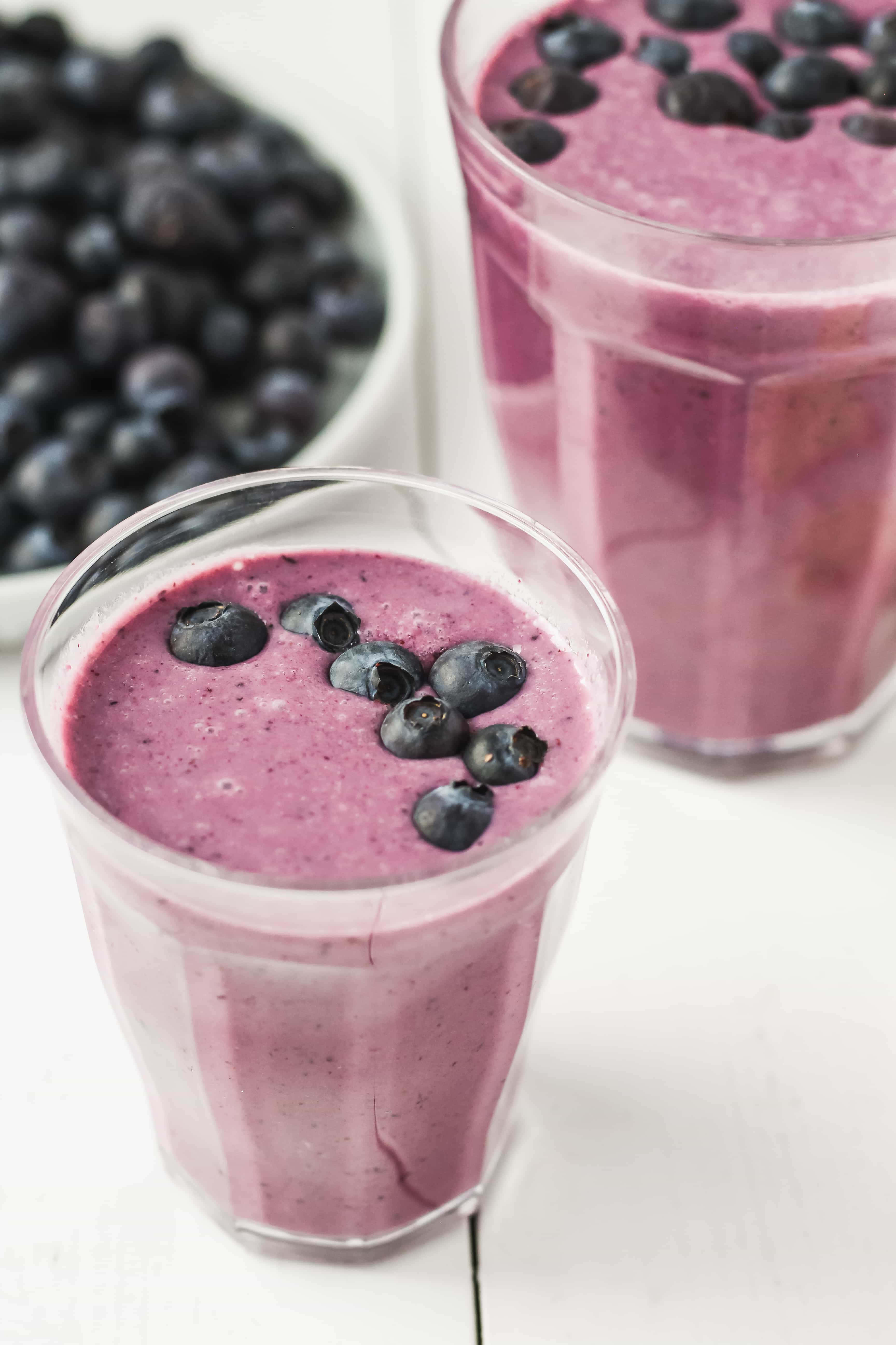Blueberry Smoothie 桌布 壁紙 — 兩杯充滿活力的紫色藍莓冰沙，上面放上新鮮藍莓，旁邊放著一碗莓果，放在白色木質表面上。