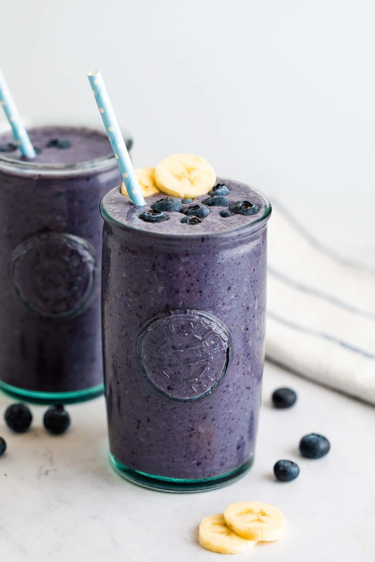 Blueberry Smoothie 桌布 壁紙 — 兩杯充滿活力的紫藍莓香蕉冰沙，裝在玻璃罐中，上面放上新鮮水果和圓點吸管。