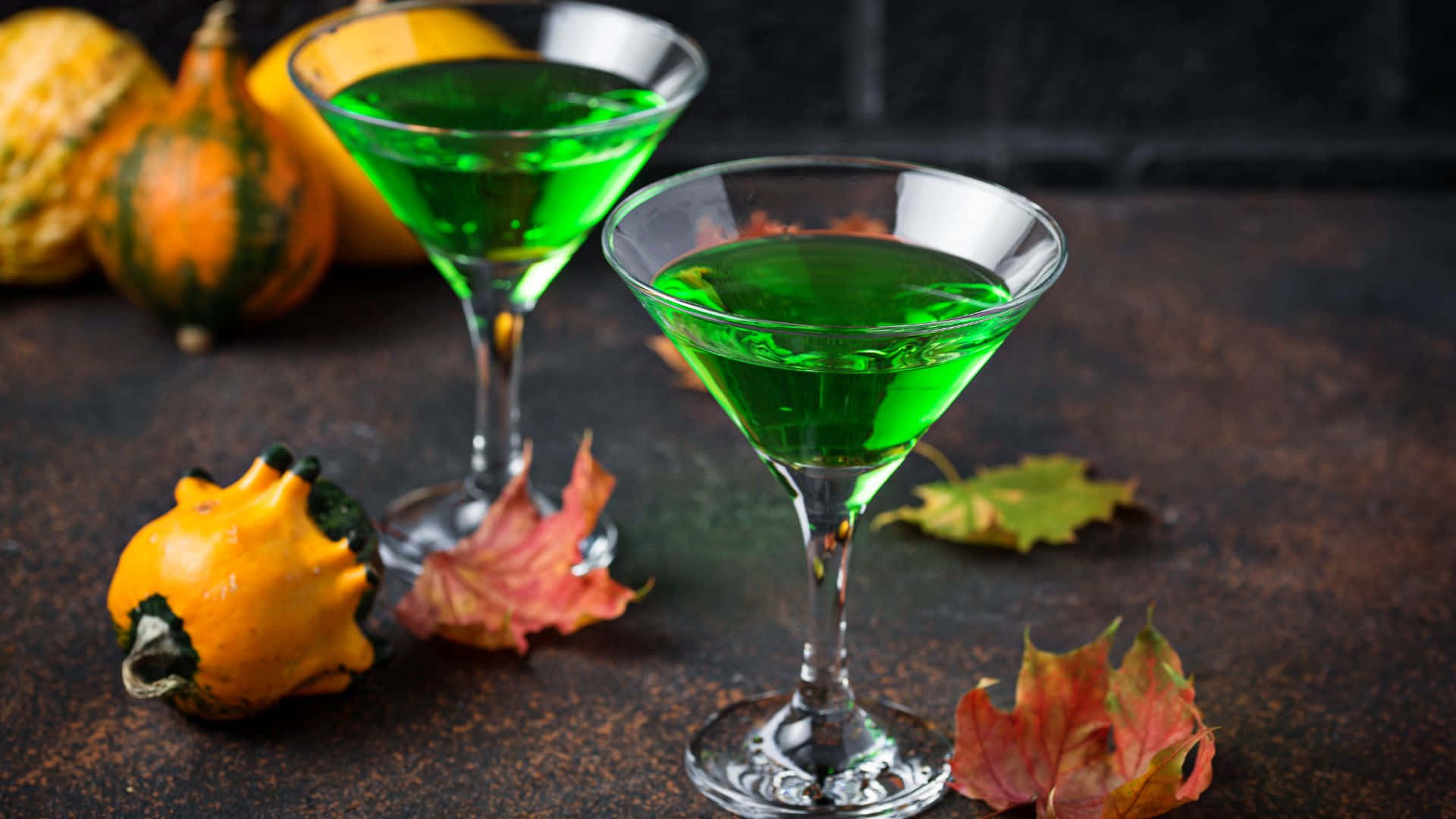 Spooky Cocktails 桌布 壁紙 — 兩杯充滿活力的綠色雞尾酒盛載於馬丁尼杯中，周圍環繞著秋季葫蘆和色彩繽紛的樹葉，置於鄉村風格的表面上。