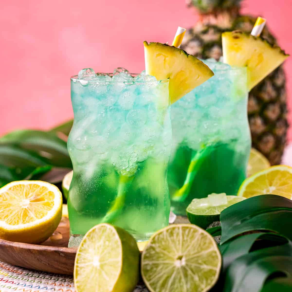 Beautiful Drink 桌布 壁紙 — 兩杯充滿活力的藍綠色熱帶雞尾酒，搭配鳳梨裝飾和柑橘片，放在粉紅色背景前的木托盤上。
