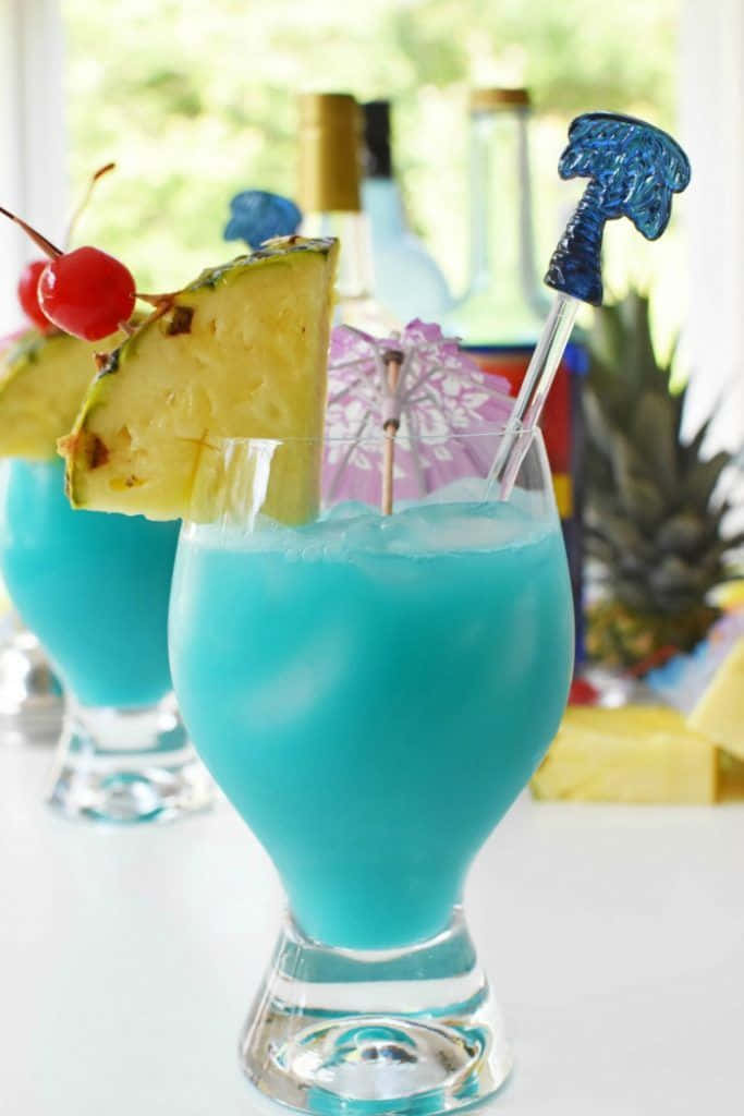 Blue Hawaii 桌布 壁紙 — 兩杯充滿活力的藍色熱帶雞尾酒，飾以鳳梨、櫻桃和棕櫚形攪拌棒，放在明亮的白色表面上。