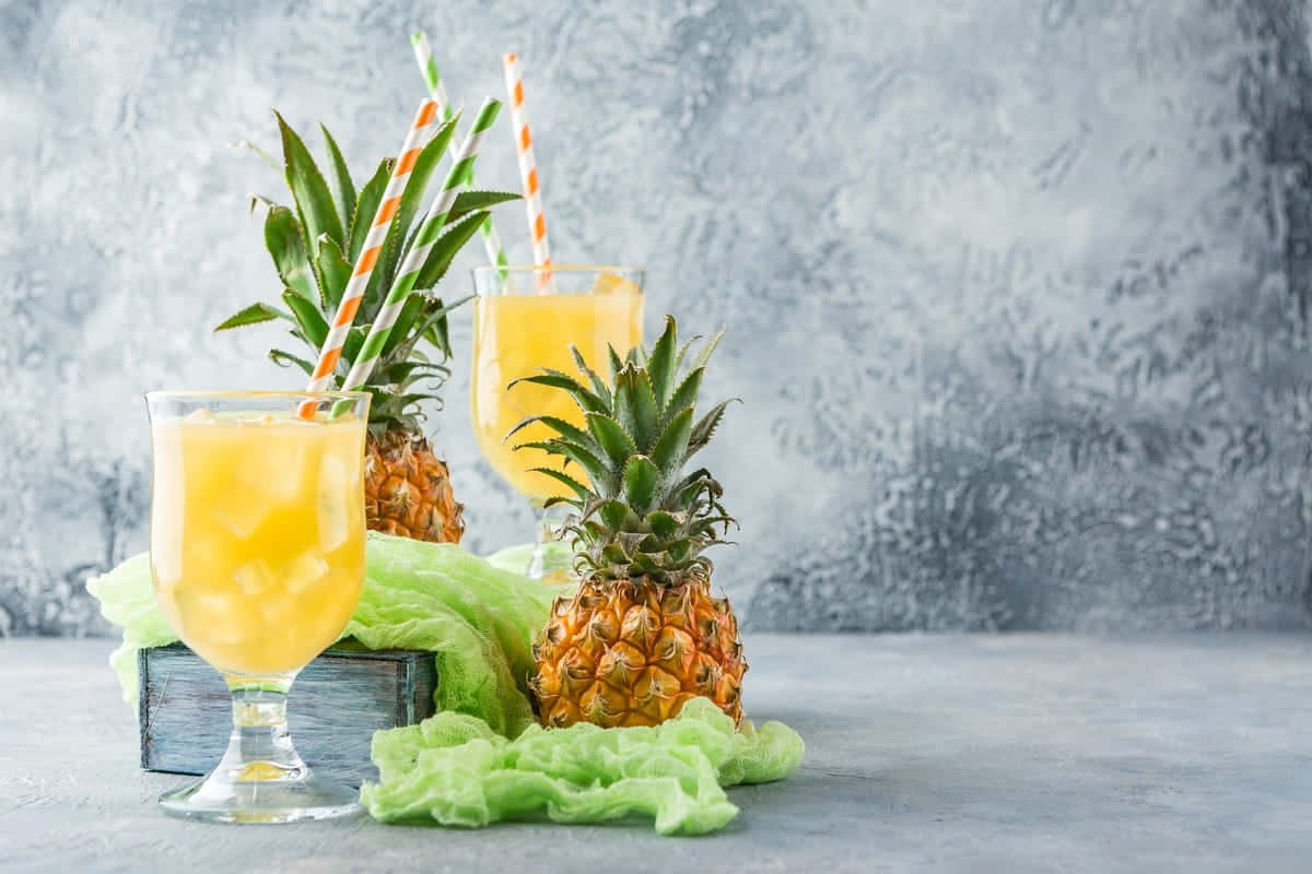 Fruit 桌布 壁紙 — 兩杯充滿活力的鳳梨汁，搭配整顆鳳梨和條紋吸管，放在綠色布上，背景為紋理灰色。