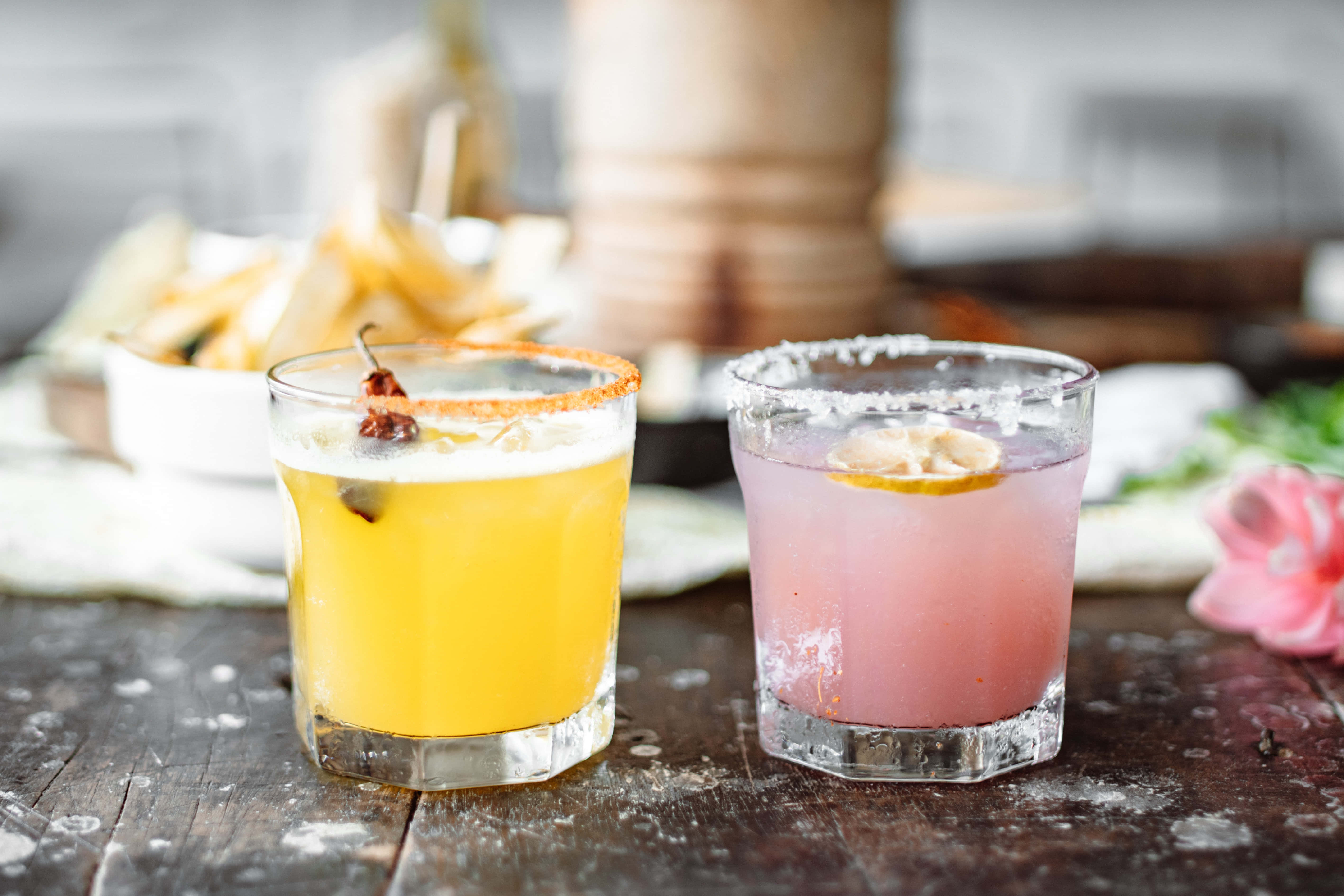 Summer Drinks 桌布 壁紙 — 兩杯充滿活力的黃色和粉紅色雞尾酒，飾以柑橘和辣椒，放在質樸的木桌上，旁邊有薯片和一朵粉紅色的花。