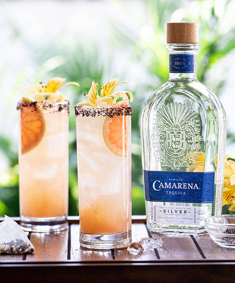Tequila 桌布 壁紙 — 兩杯充滿活力的龍舌蘭雞尾酒，配上柑橘和花卉裝飾，放在木桌上，旁邊是一瓶Camarena Silver龍舌蘭酒。