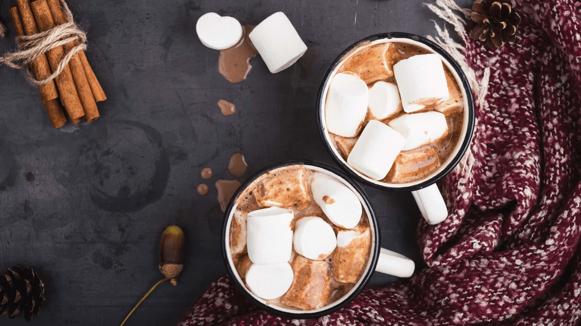Hot Chocolate 桌布 壁紙 — 兩杯冒著熱氣的熱巧克力，上面放著棉花糖，旁邊放著肉桂棒、松果和一條舒適的圍巾，放在深色紋理表面上。