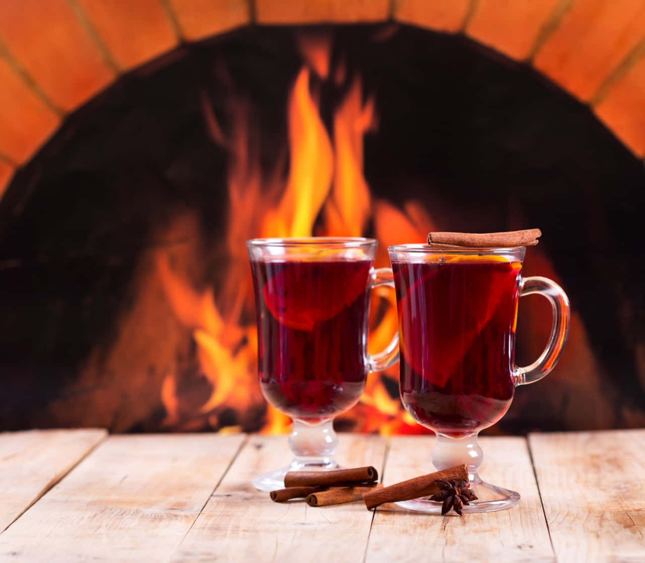 Winter Drinks 桌布 壁紙 — 兩杯冒著熱氣的香料熱紅酒，配上肉桂棒和八角，放在木桌上，背景是噼啪作響的磚砌壁爐。