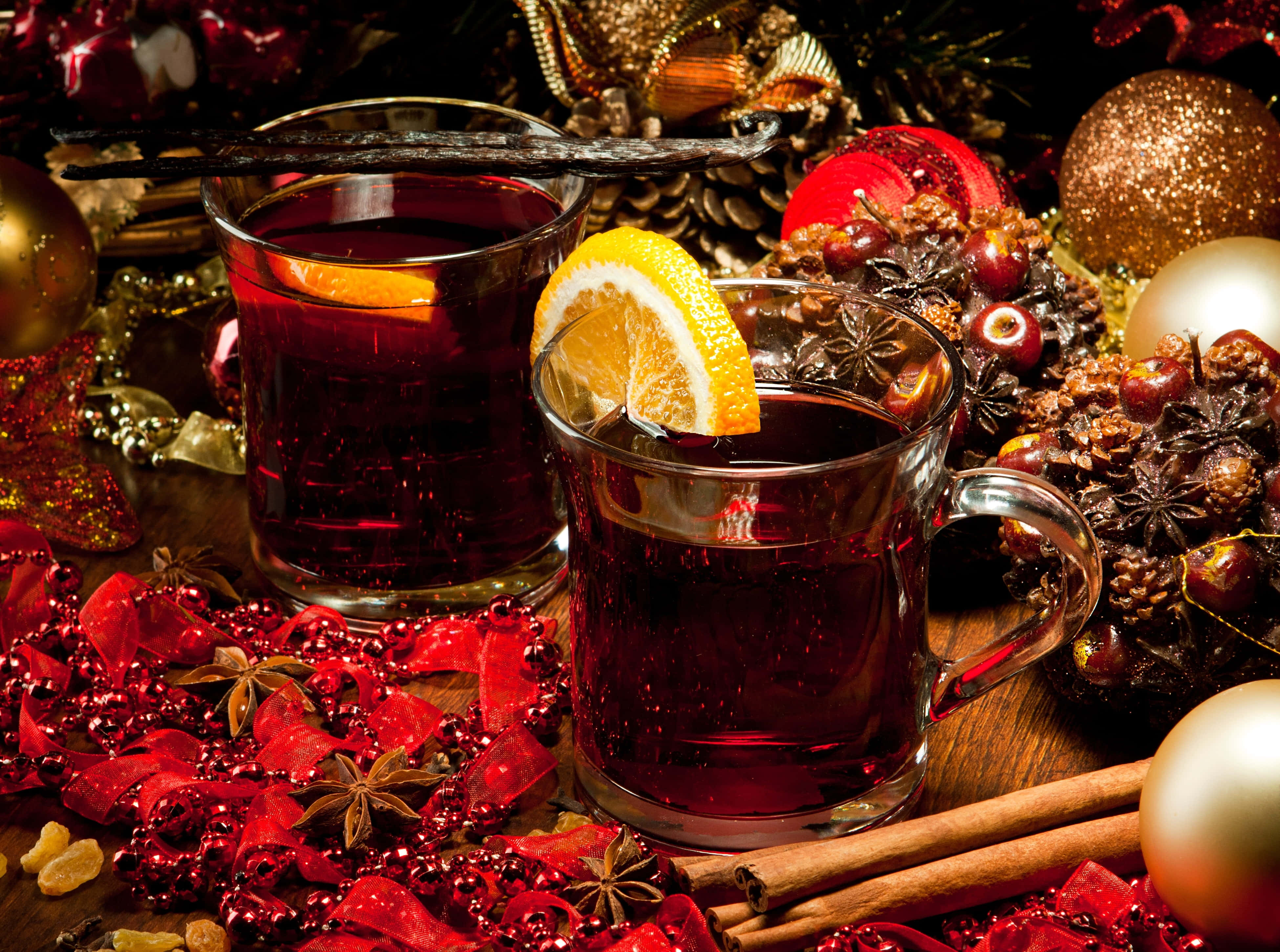 Mulled Wine 桌布 壁紙 — 兩杯冒著熱氣的香料熱紅酒，飾以柑橘和香草，放在木質表面上，周圍是節日紅色裝飾品、松果和香料。