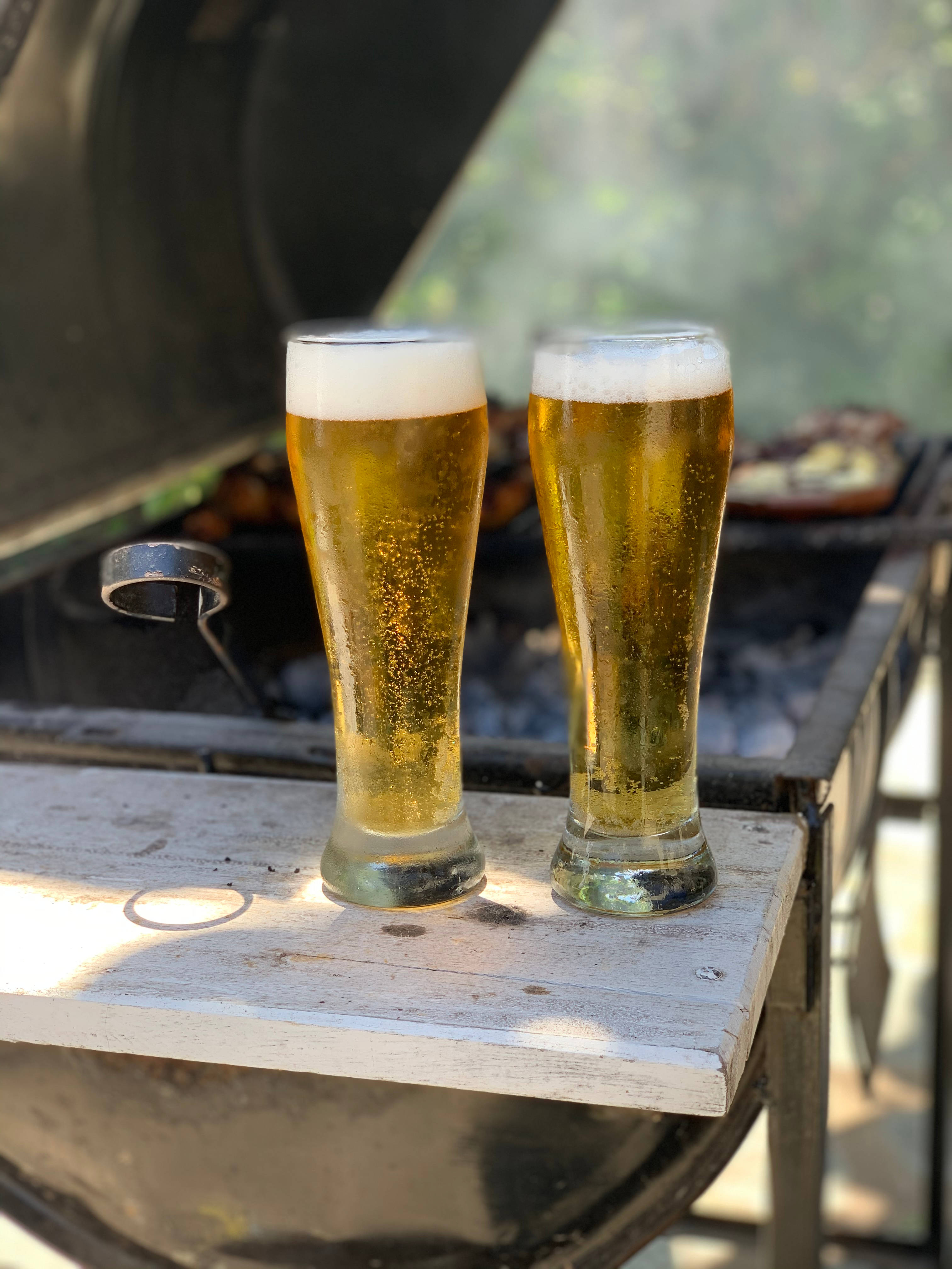 Beer 桌布 壁紙 — 兩杯冰凍的皮爾森啤酒杯盛著金黃色啤酒和白色泡沫，放在一張風化的木桌上，旁邊有一個開放式烤架，食物正在滋滋作響。
