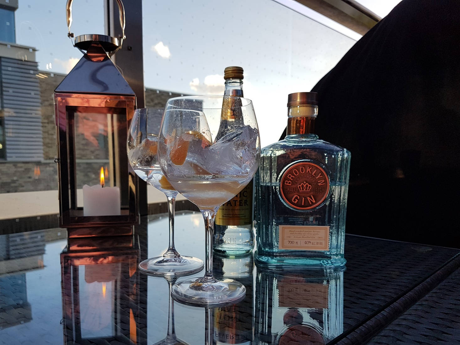 alcohol 桌布 壁紙 — 兩杯加冰和橙片的琴酒雞尾酒，放在日落時分的反光桌上，旁邊有一盞點亮的燈籠和琴酒瓶。