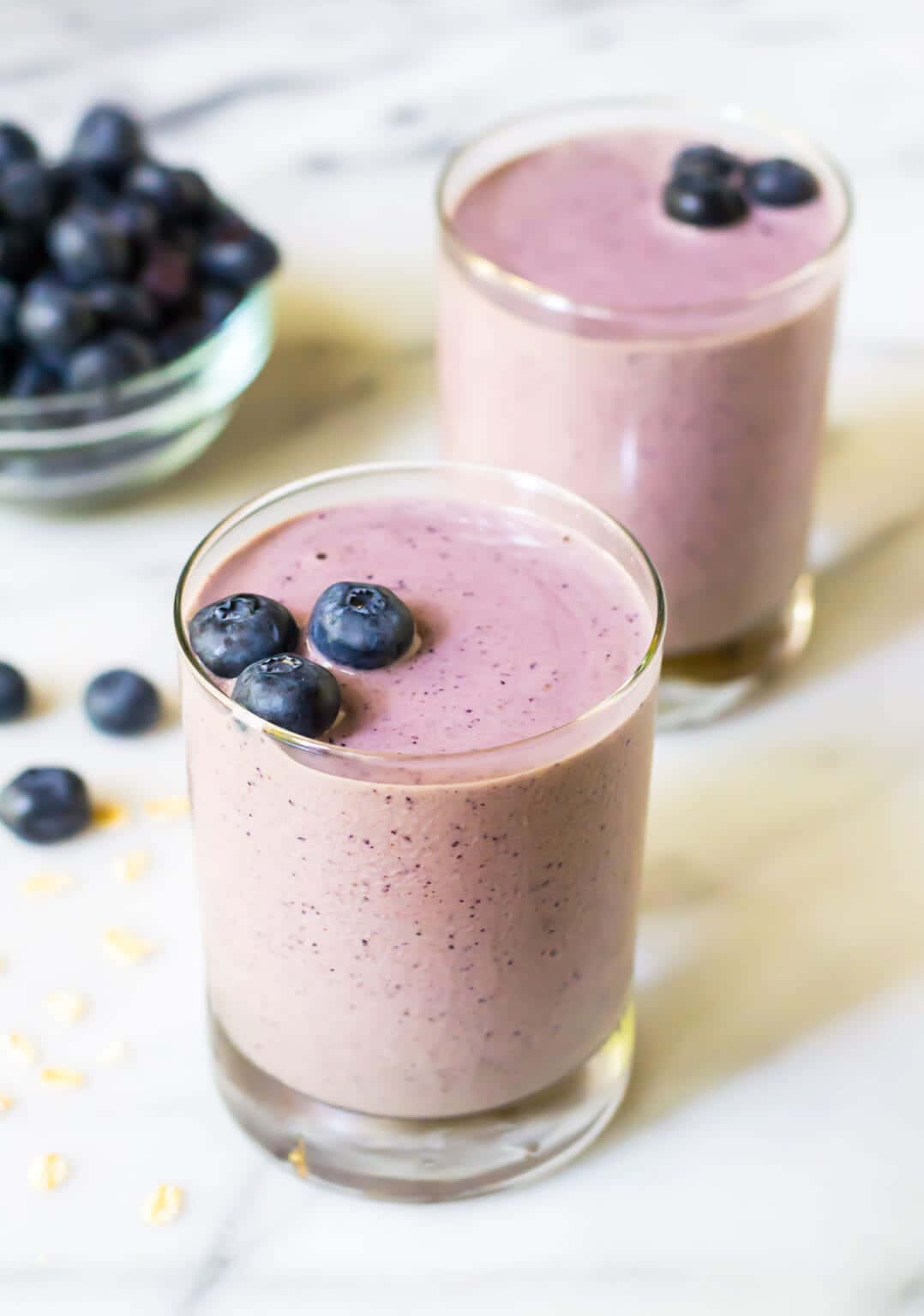 Blueberry Smoothie 桌布 壁紙 — 兩杯奶油紫色藍莓冰沙，上面放上新鮮藍莓，旁邊有一碗莓果和散落的燕麥片，放在大理石表面上。