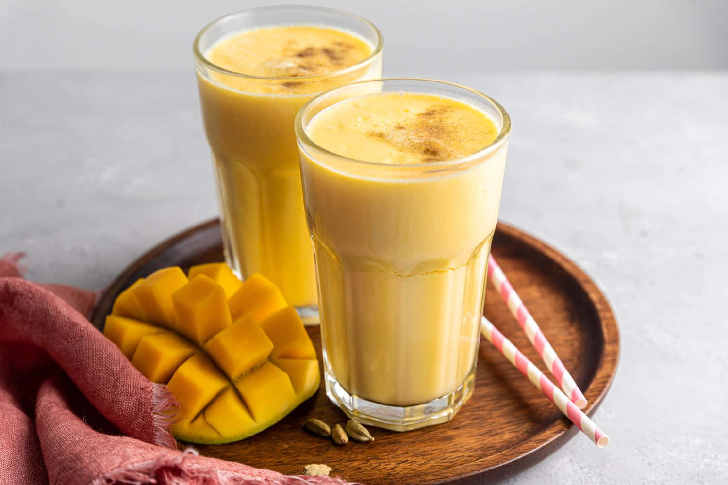 Mango 桌布 壁紙 — 兩杯奶油芒果拉西，上面撒有肉桂粉，放在木托盤上，旁邊是新鮮芒果丁和粉紅條紋吸管。