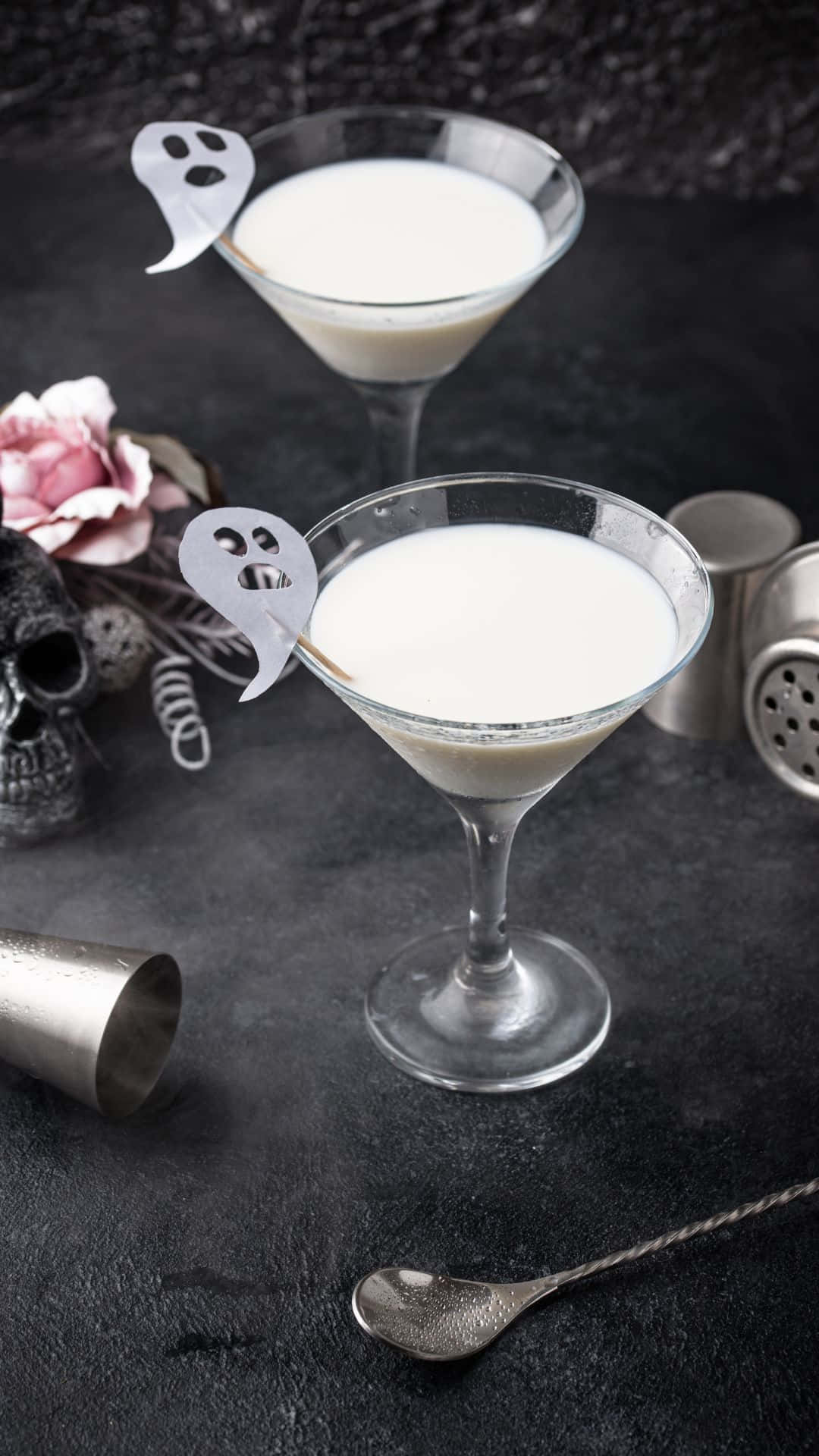 Spooky Cocktails 桌布 壁紙 — 兩杯奶油雞尾酒放在馬丁尼杯中，飾有幽靈攪拌棒，旁邊有一個骷髏頭和一朵粉紅玫瑰，置於深色紋理表面上。