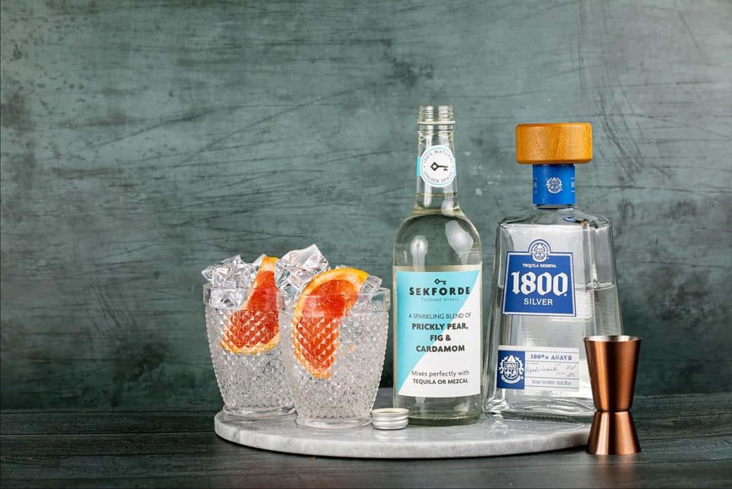 alcohol 桌布 壁紙 — 兩杯帶有仙人掌梨裝飾的雞尾酒，旁邊放著 Sekforde 和 1800 Silver 龍舌蘭酒瓶，放在大理石托盤上，背景是紋理灰色牆壁。
