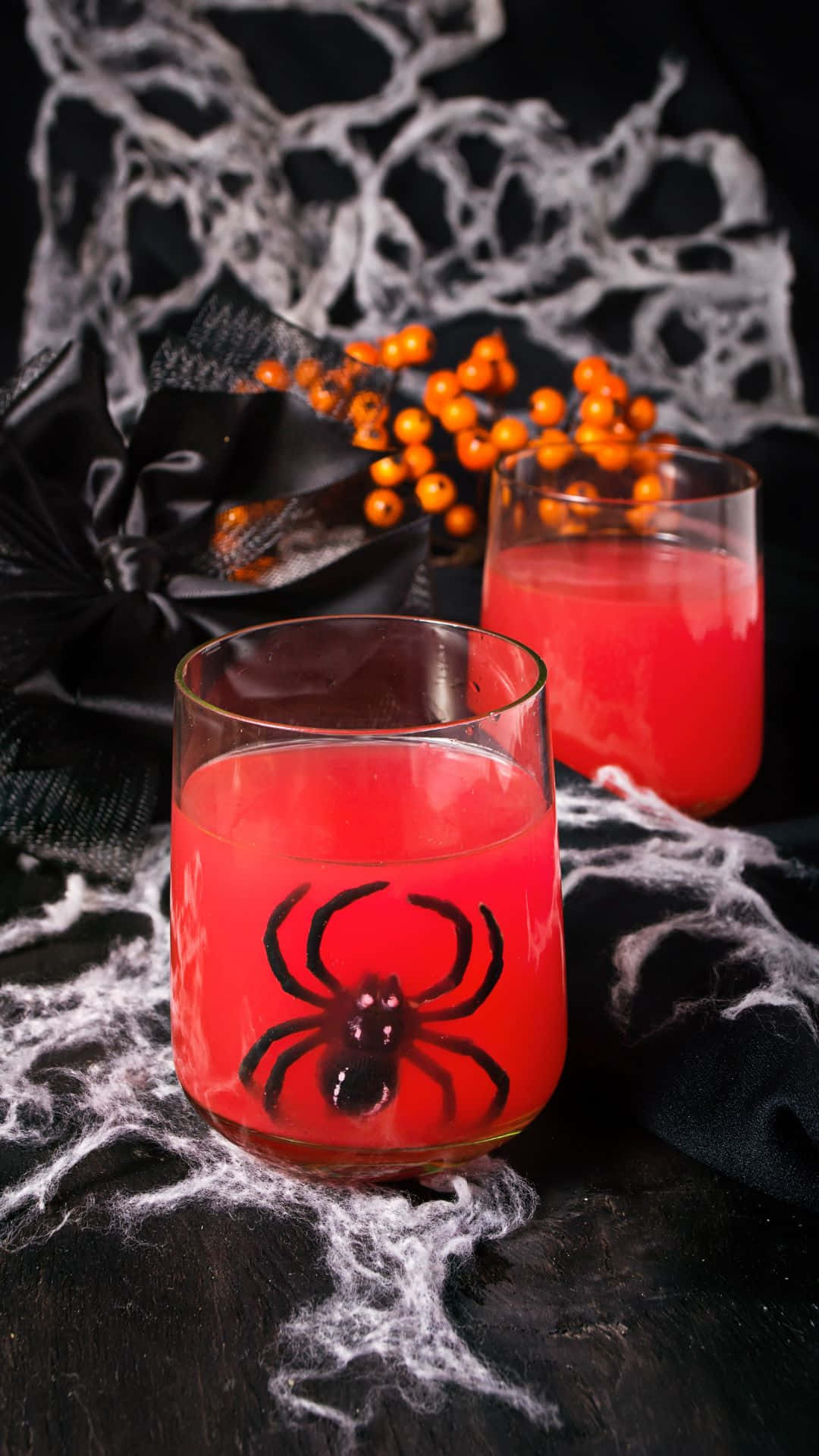 Spooky Cocktails 桌布 壁紙 — 兩杯帶有蜘蛛貼紙的紅色萬聖節潘趣酒，放在鋪有白色蜘蛛網和橙色漿果的黑色表面上。