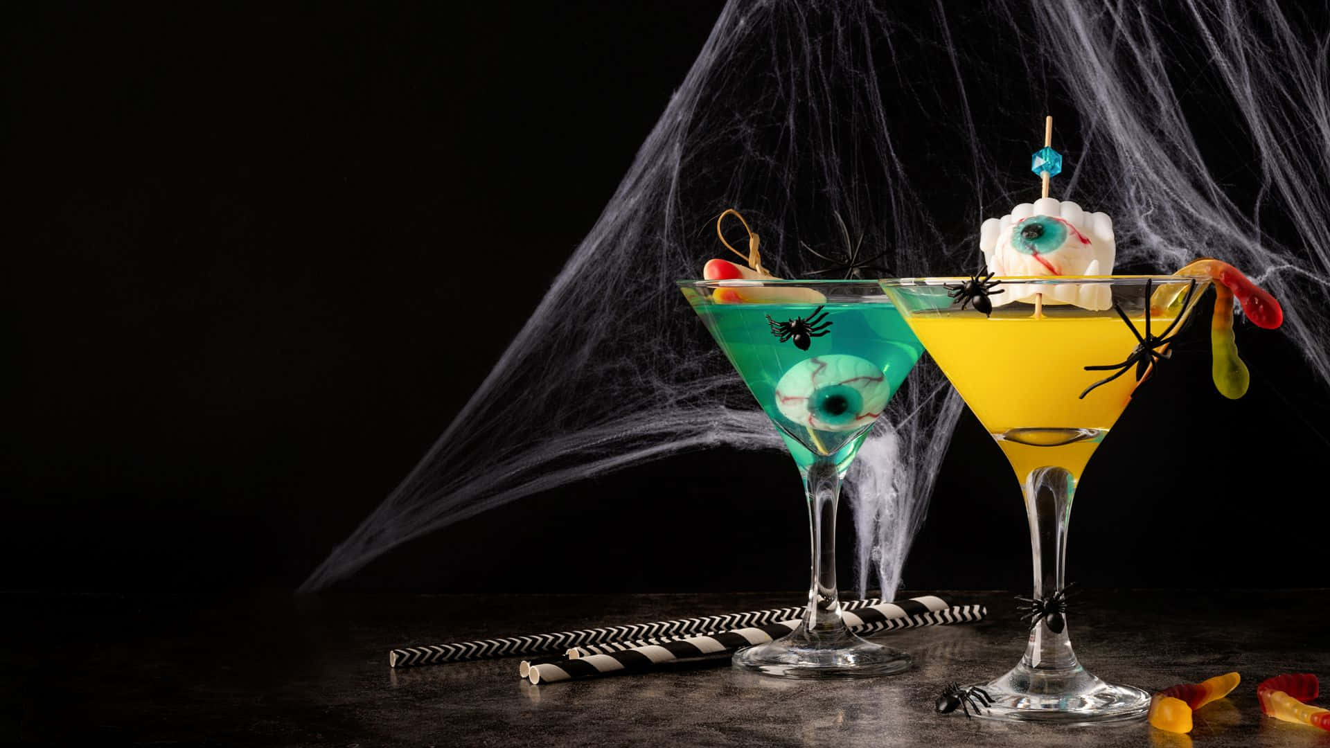 Spooky Cocktails 桌布 壁紙 — 兩杯恐怖萬聖節雞尾酒，盛載於馬丁尼杯中，飾以眼珠和蜘蛛，背景為黑色帶蜘蛛網。