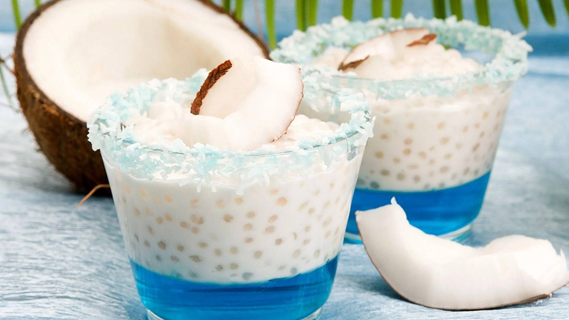 Coconut 桌布 壁紙 — 兩杯椰子西米露甜品，搭配藍色糖漿和椰子片裝飾，旁邊有一個裂開的椰子，置於藍色紋理表面上。