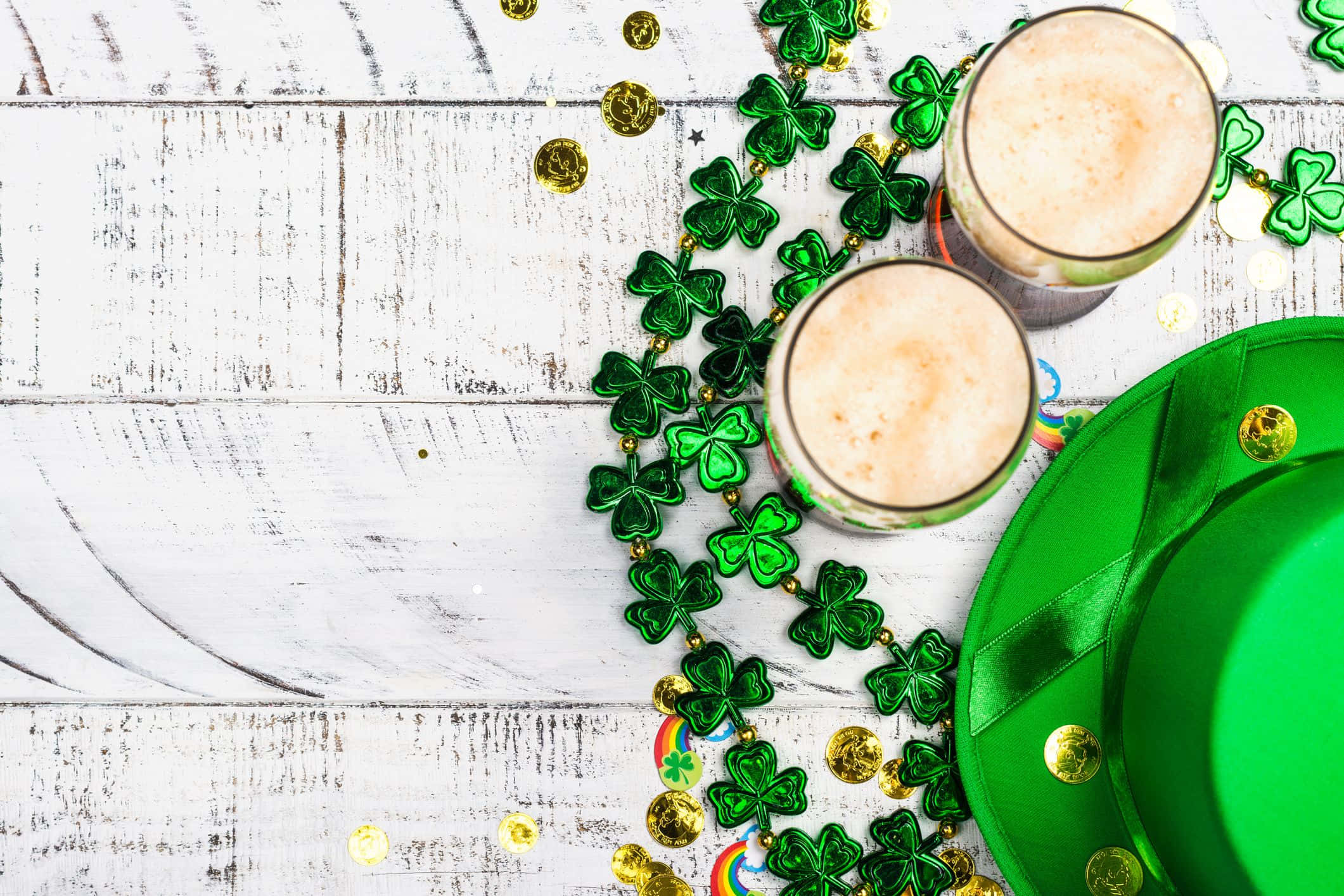 St Patrick's Day Zoom 桌布 壁紙 — 兩杯泡沫啤酒和一頂綠色帽子，被三葉草珠子和金幣包圍，放在白色木質表面上。