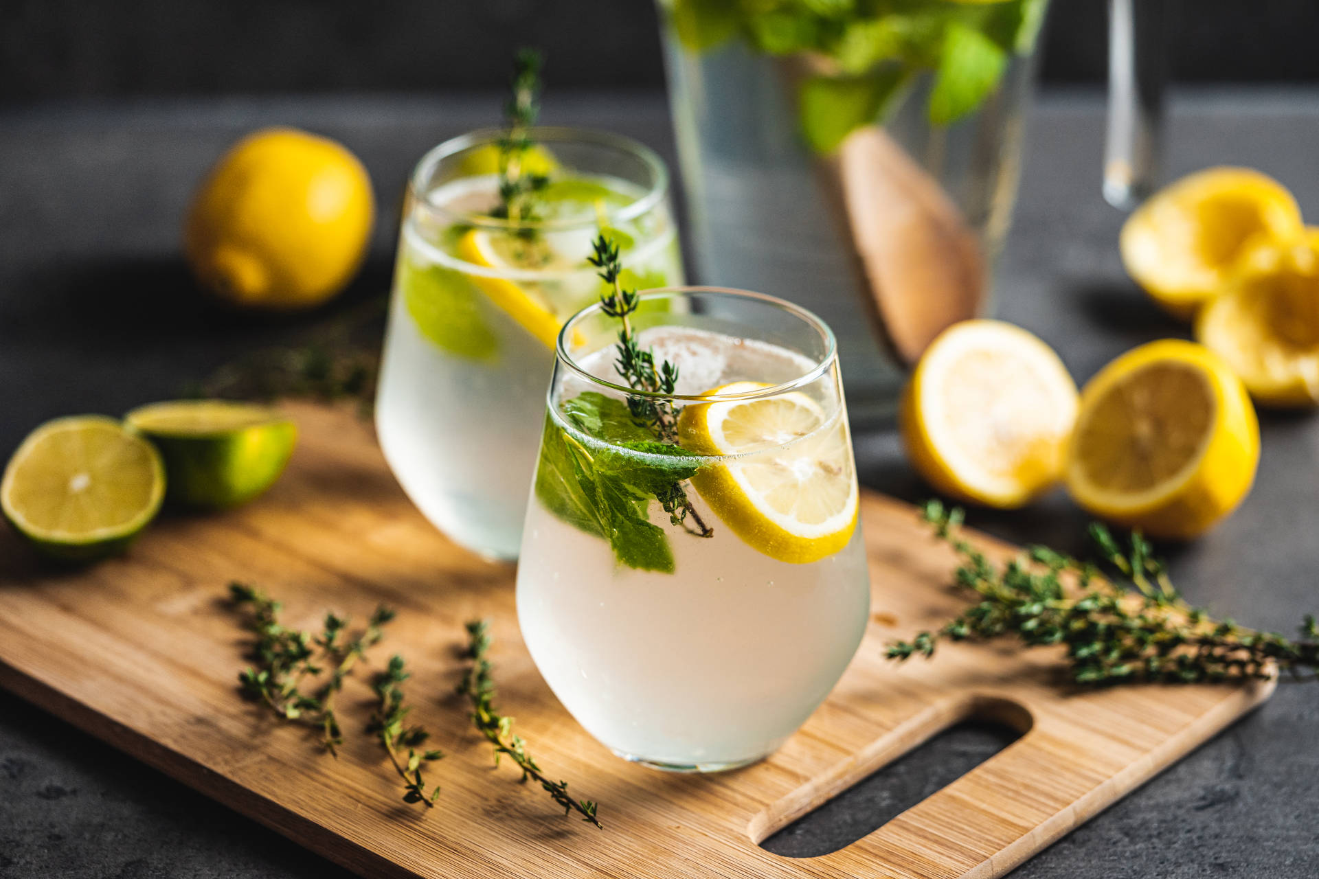 Lemon 桌布 壁紙 — 兩杯清爽的檸檬百里香飲品，飾以柑橘片和香草，放在一個木製砧板上，周圍是新鮮的檸檬和萊姆。