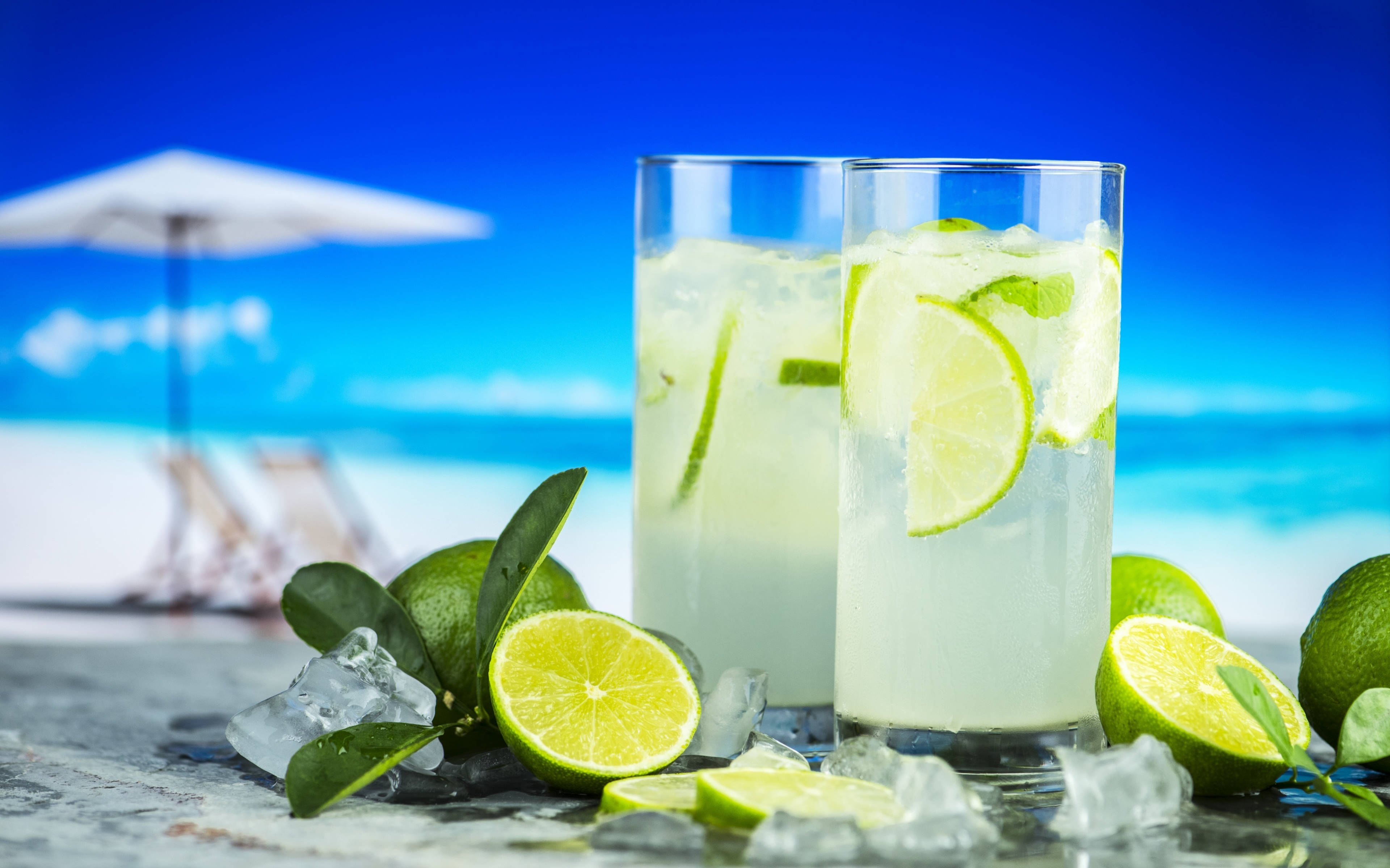 4K Summer 桌布 壁紙 — 兩杯清爽的萊姆雞尾酒，加冰和裝飾，放在石頭表面上，背景是充滿活力的藍色海灘和白色遮陽傘。