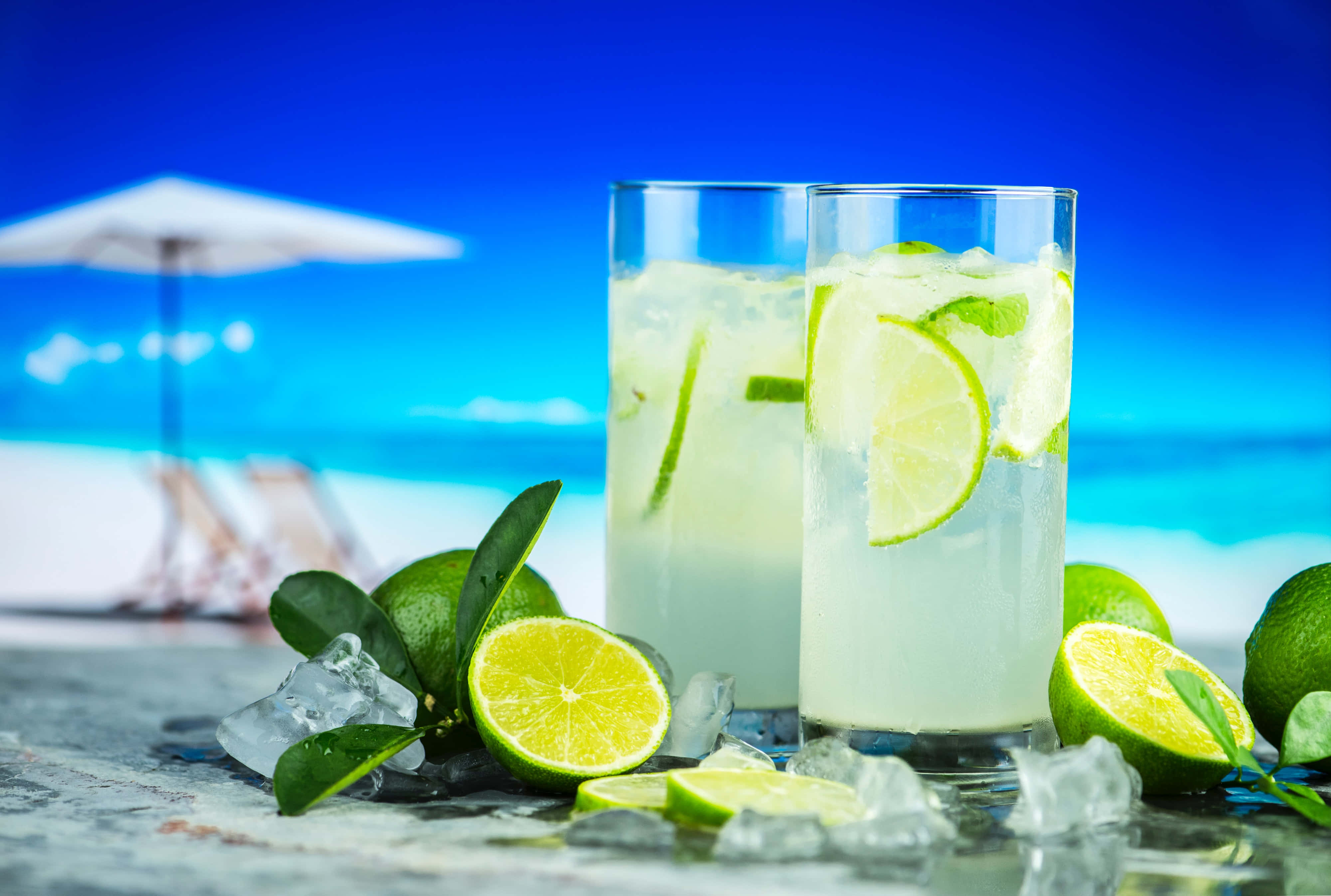 Hydration 桌布 壁紙 — 兩杯清爽的青檸雞尾酒配上冰塊和裝飾，放在桌子上，背景是充滿活力的藍色海灘和白色遮陽傘。