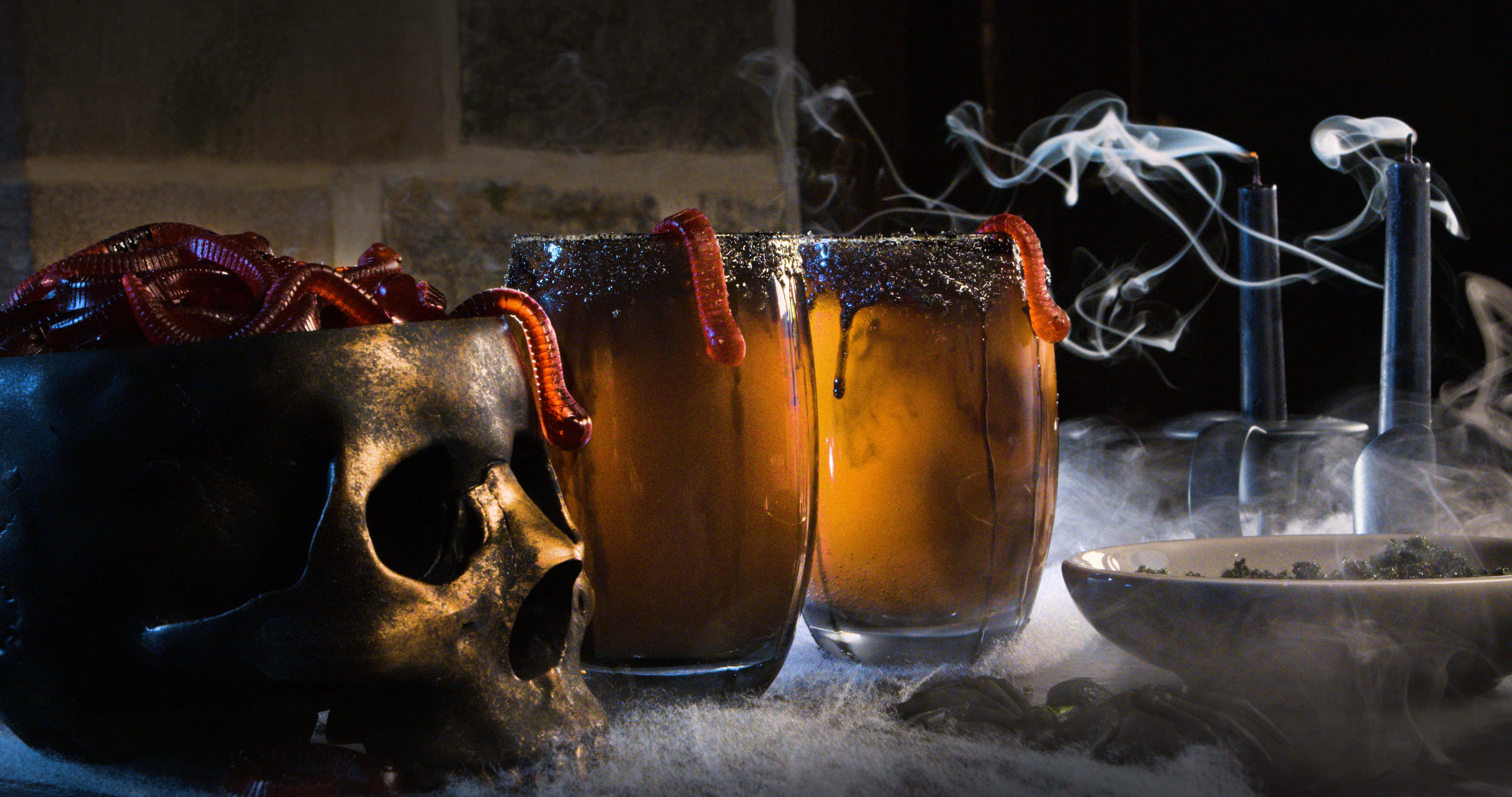 Spooky Cocktails 桌布 壁紙 — 兩杯煙燻琥珀色飲料，內有蠕動的蟲子，旁邊放著一個骷髏頭和冒煙的煙斗，置於結霜的表面上。