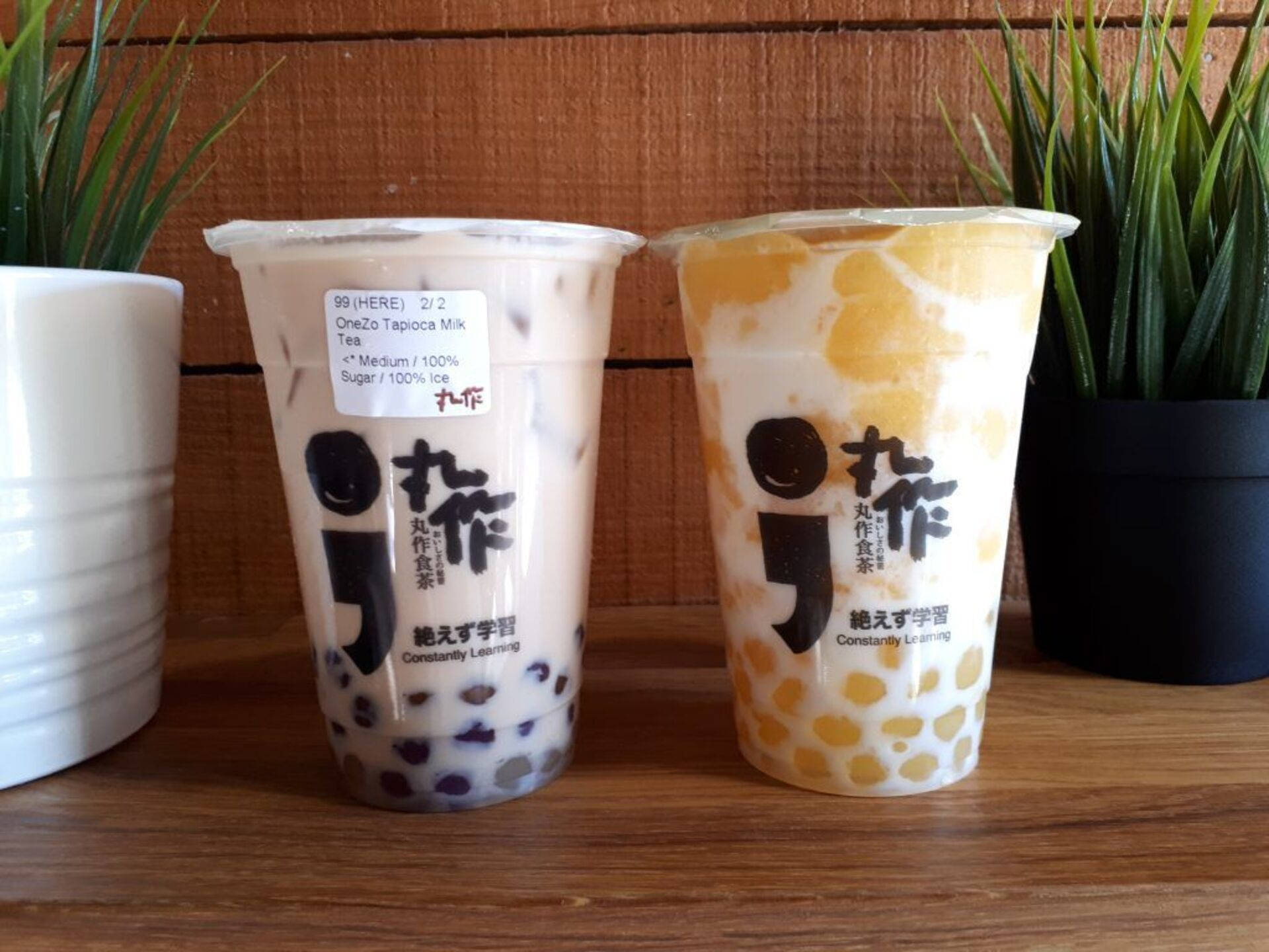 Bubble 桌布 壁紙 — 兩杯珍珠奶茶，含有木薯珍珠和芒果珍珠，放在一張木桌上，兩旁有盆栽植物。