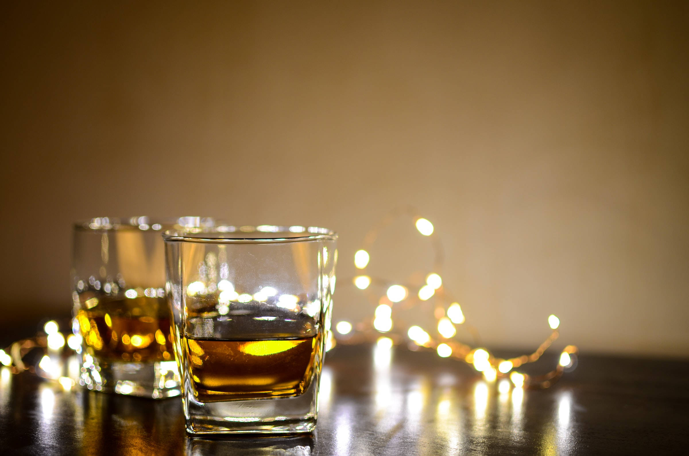 Drink 桌布 壁紙 — 兩杯琥珀色威士忌在深色桌子上溫暖地發光，旁邊閃爍著仙女燈，背景是柔和的米色。