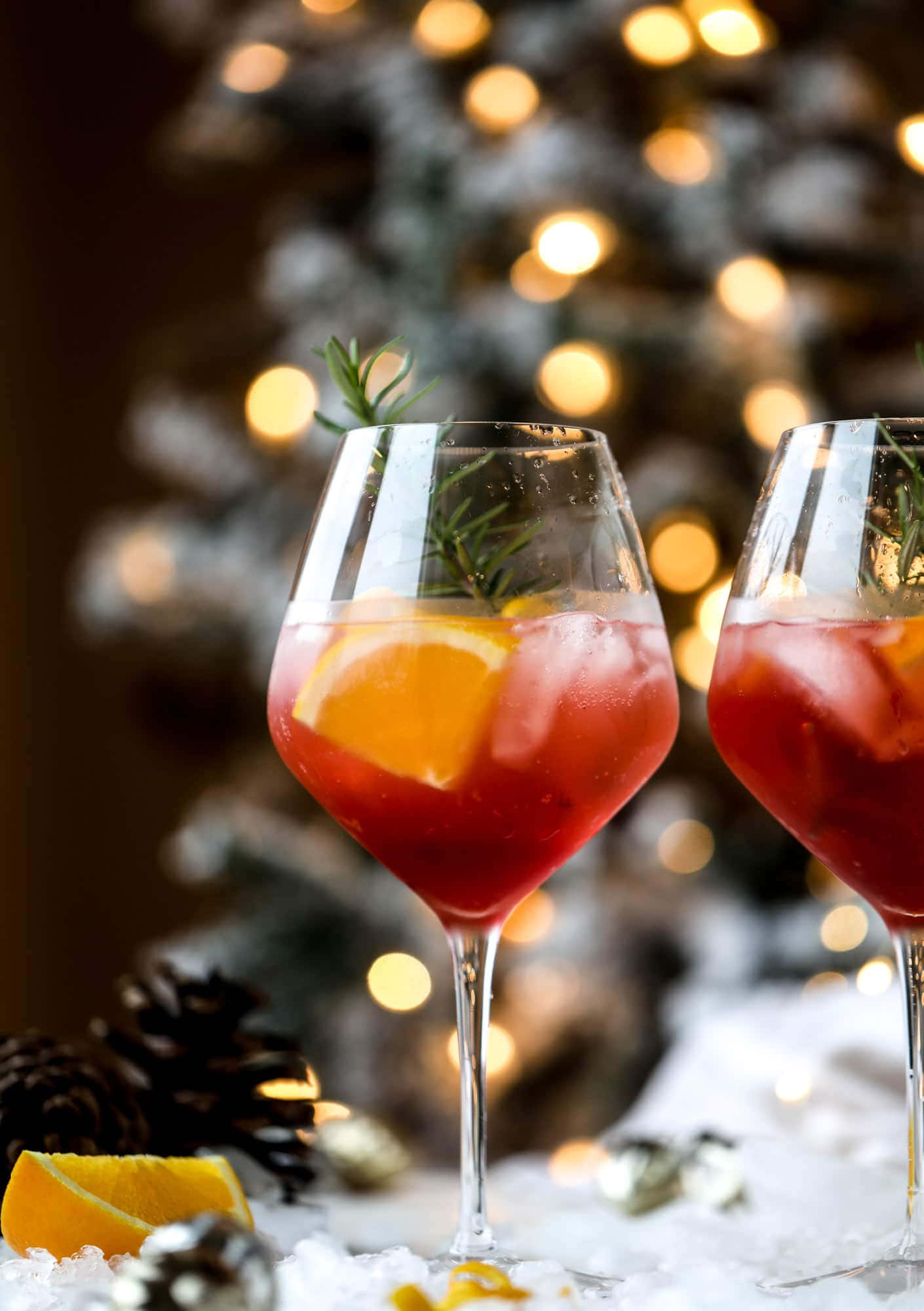 Winter Drinks 桌布 壁紙 — 兩杯節日紅色雞尾酒，飾以橙片和迷迭香，放在雪地上，背景是模糊的聖誕樹燈光。