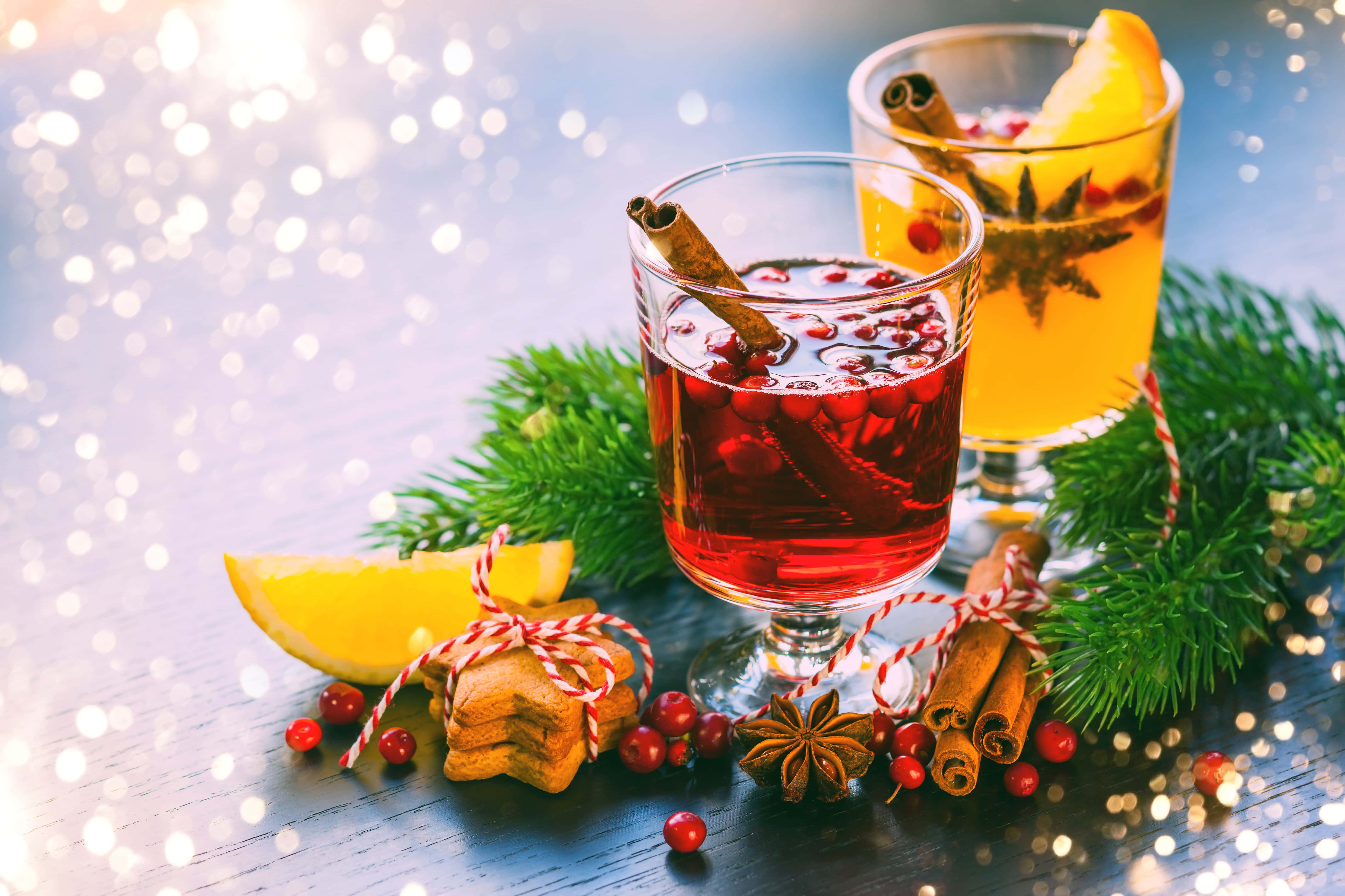 Mulled Wine 桌布 壁紙 — 兩杯節日飲品，以肉桂棒、八角茴香和柑橘裝飾，旁邊放著松枝和紅莓，置於深色木桌上，背景有閃爍的散景燈光。