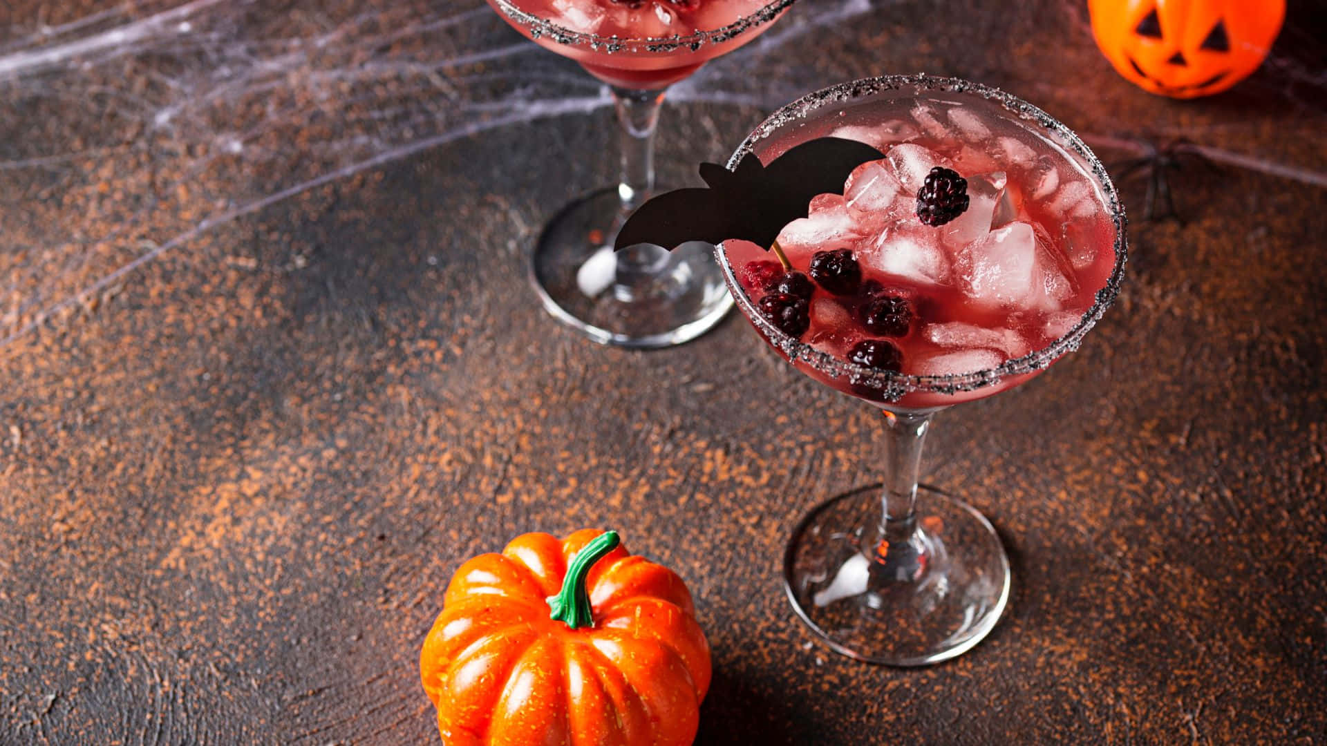 Halloween Cocktails 桌布 壁紙 — 兩杯粉紅色萬聖節雞尾酒，飾有黑莓和蝙蝠裝飾，放在深色紋理表面的南瓜旁邊。