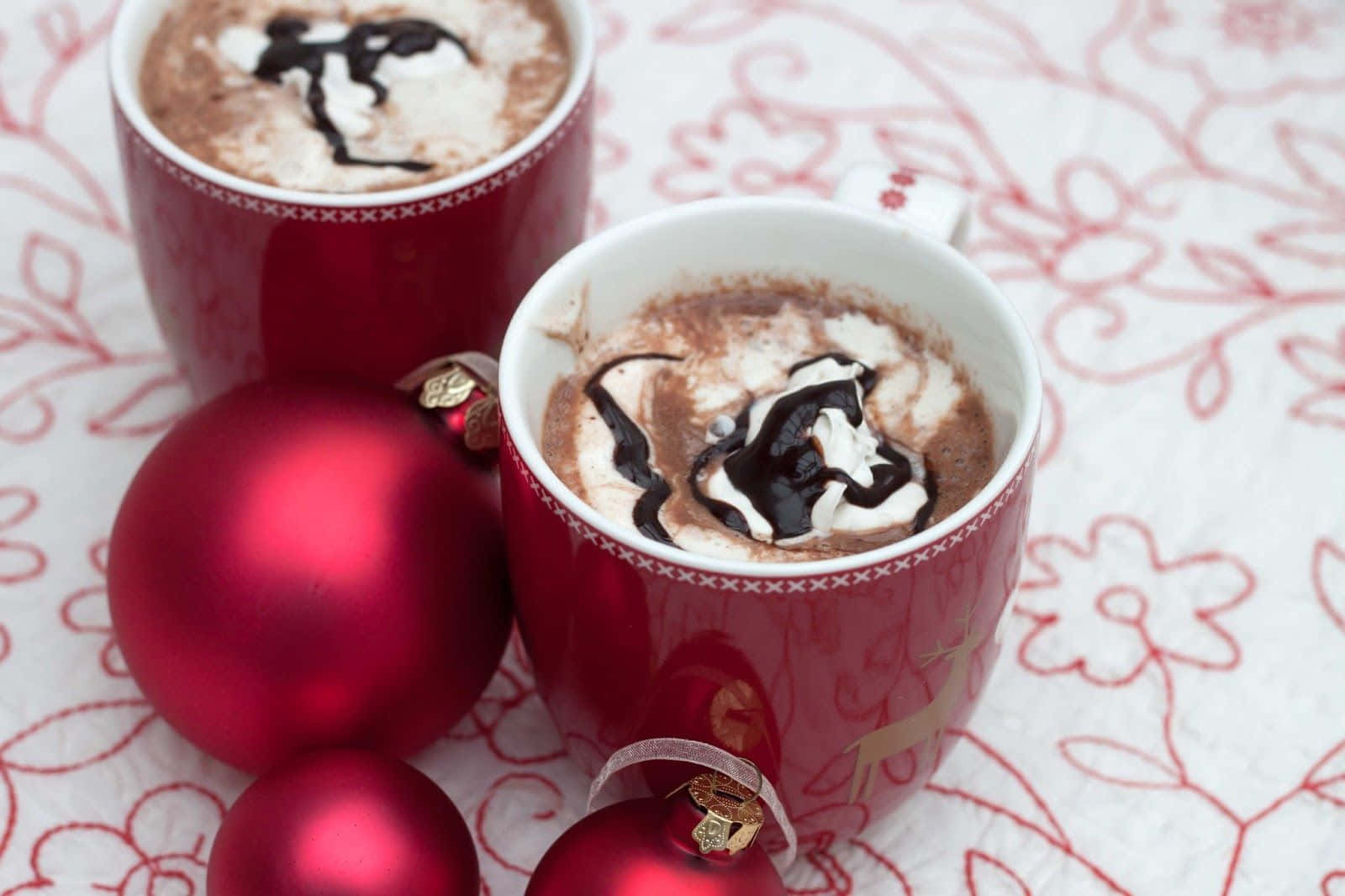 Hot Chocolate 桌布 壁紙 — 兩杯紅色馬克杯的熱巧克力，配上鮮奶油和巧克力醬，旁邊放著閃亮的紅色裝飾品，置於花卉圖案的桌布上。