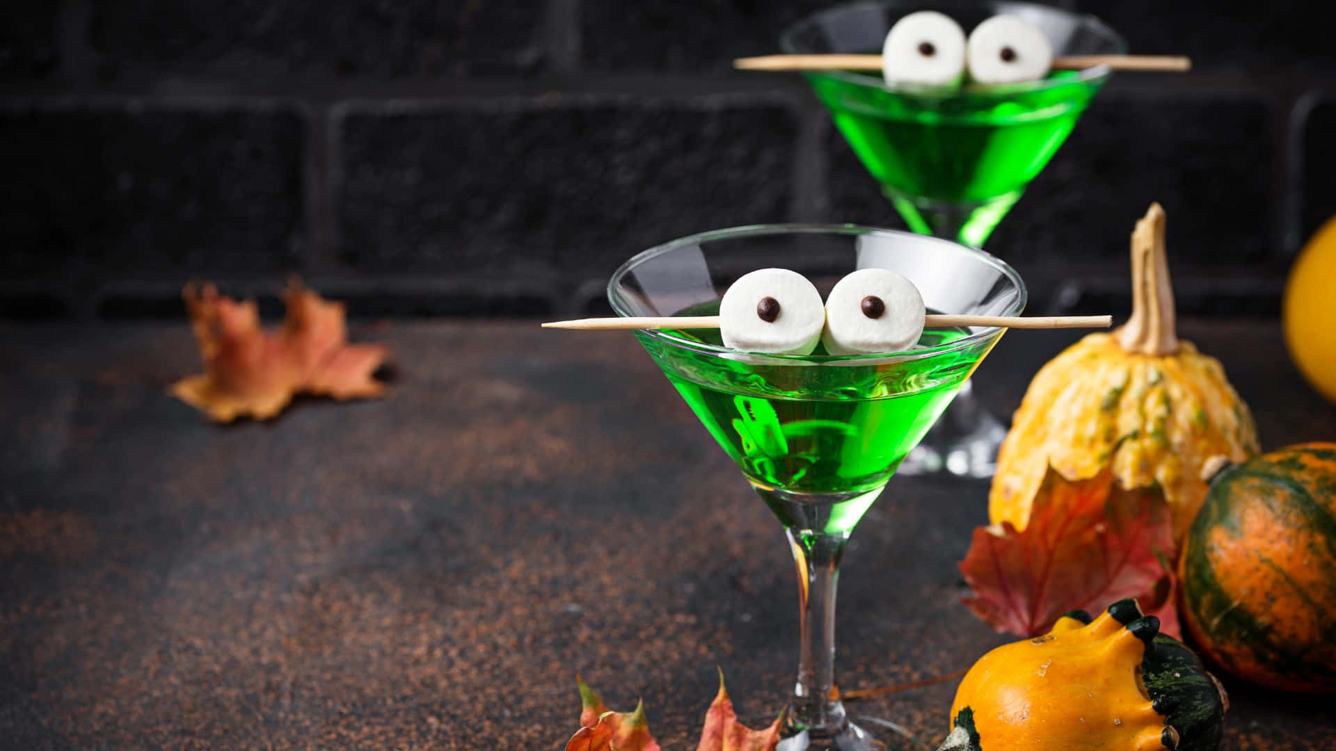 Spooky Cocktails 桌布 壁紙 — 兩杯綠色萬聖節雞尾酒，帶有棉花糖眼睛，放在深色磚牆背景上，旁邊有南瓜和秋葉。
