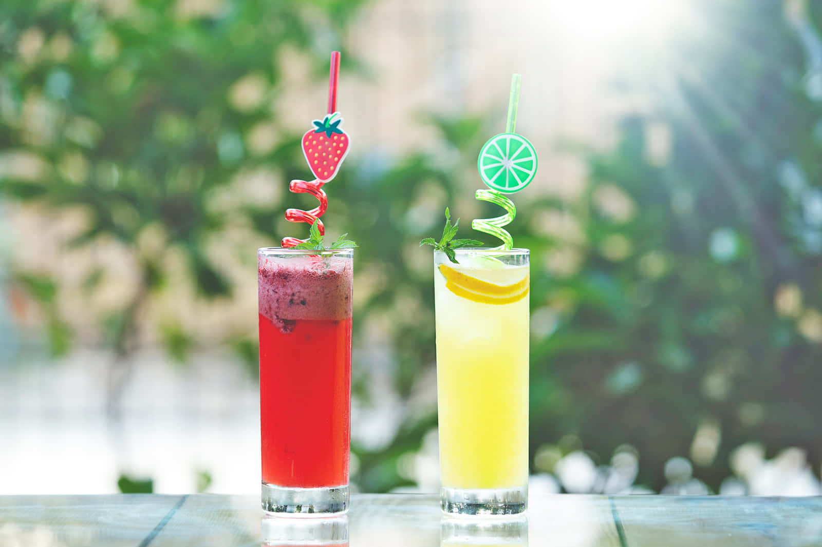 drinks 桌布 壁紙 — 兩杯色彩鮮豔的紅色和黃色雞尾酒，飾以水果和螺旋吸管，放在木桌上，背景是陽光普照的綠色。