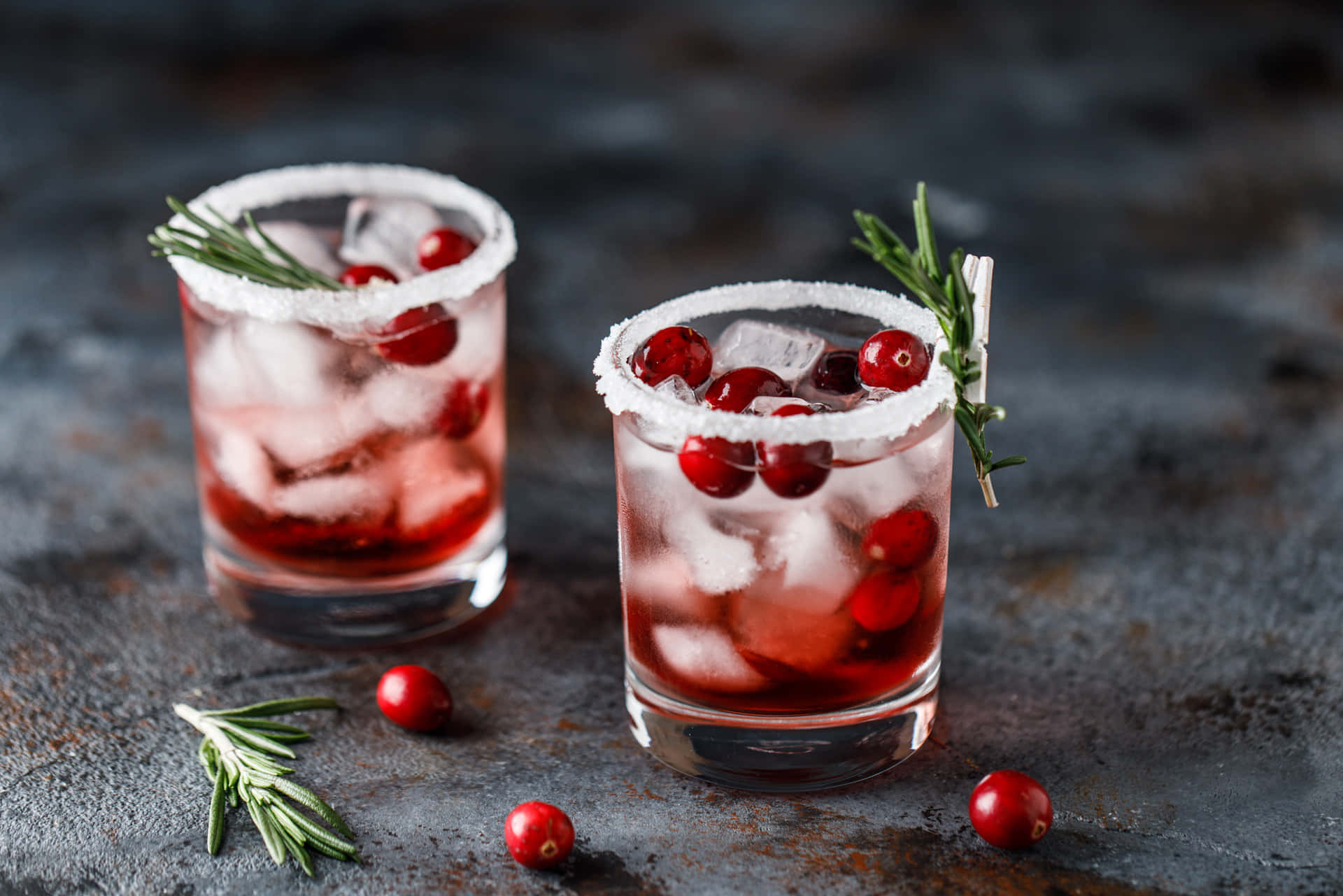 Winter Drinks 桌布 壁紙 — 兩杯蔓越莓雞尾酒，配有冰塊、迷迭香和鹽邊，放在深色紋理表面上。