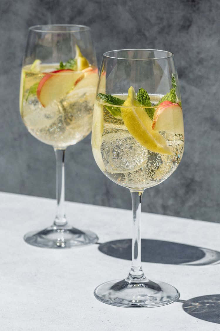 White Wine 桌布 壁紙 — 兩杯閃閃發光的琴酒雞尾酒，飾以桃子、檸檬和薄荷，放在白色表面上，背景為灰色。