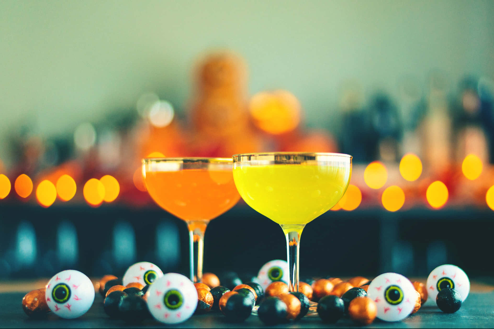 Spooky Cocktails 桌布 壁紙 — 兩杯鮮豔的橙色和黃色雞尾酒放在糖果眼珠和巧克力旁邊，背景是模糊的萬聖節主題酒吧。