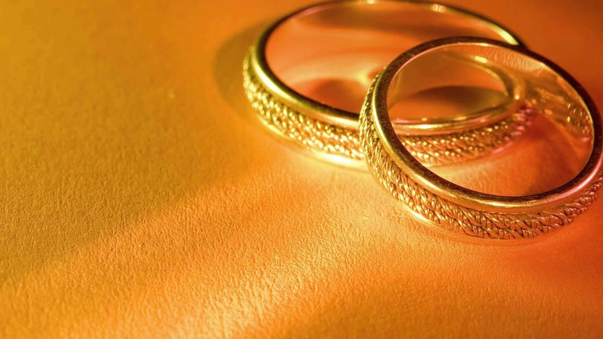 Wedding 桌布 壁紙 — 兩枚交織的金色結婚戒指在柔和的定向燈光下，在帶紋理的金色表面上溫暖地閃耀。
