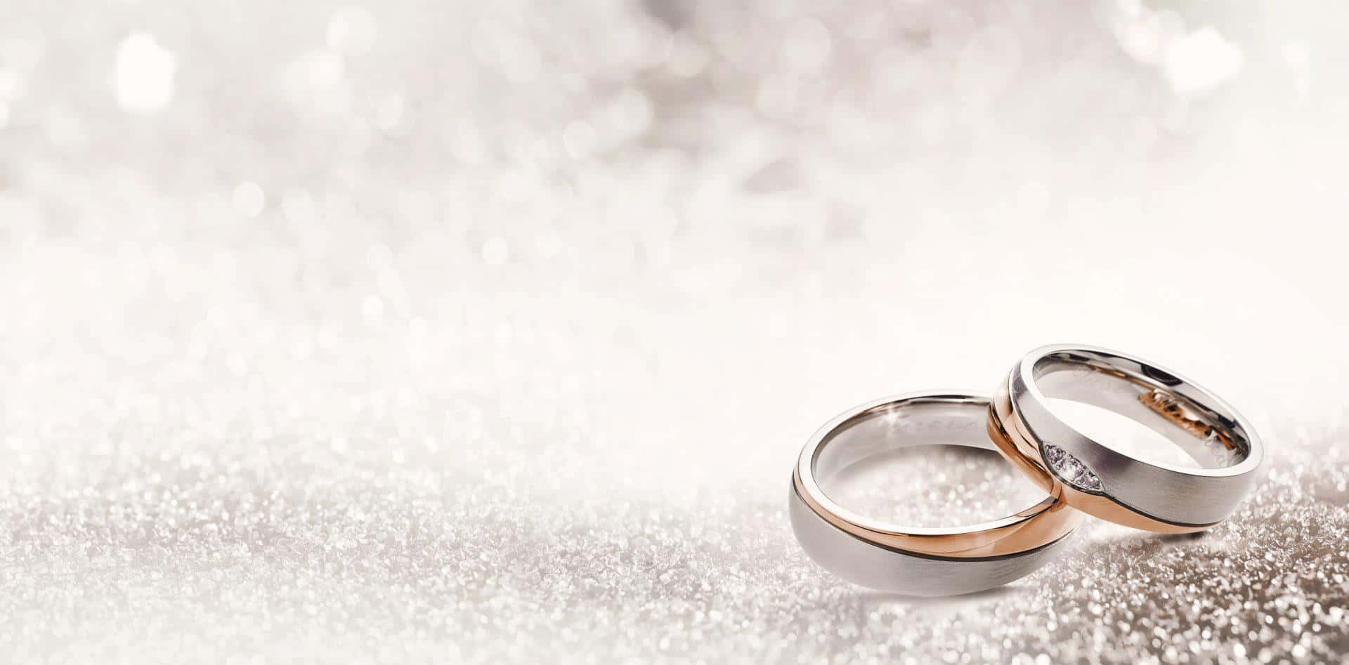結婚戒指 桌布 壁紙 — 兩枚優雅的結婚戒指，一枚玫瑰金一枚銀色，放置在閃閃發光的銀色亮粉表面上，背景柔和散景。
