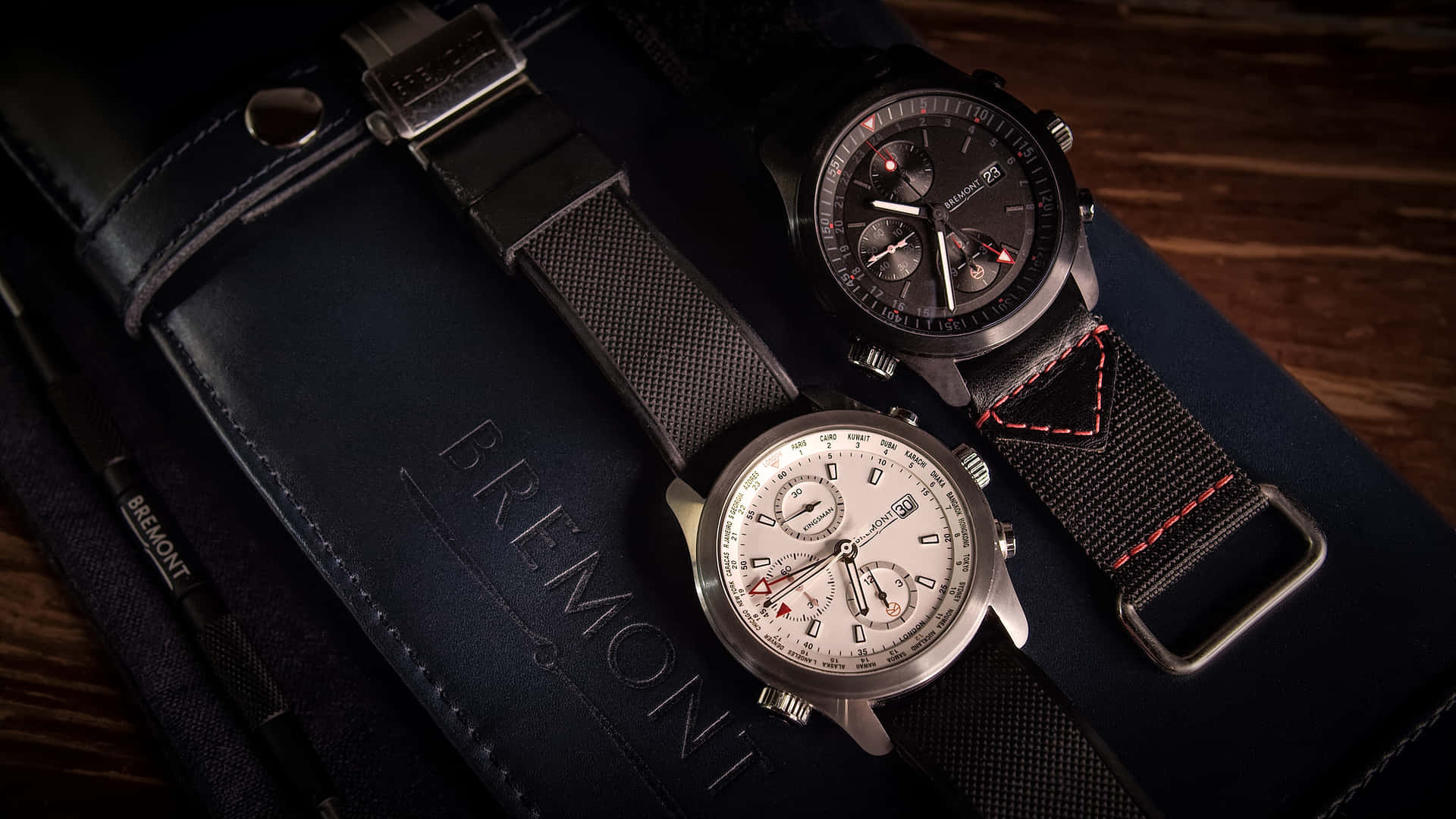 Bremont Kingsman Edition Watches 壁紙 — 兩枚帶有紋理錶帶的Bremont計時碼錶放在木質表面上的海軍藍皮革錶盒上。