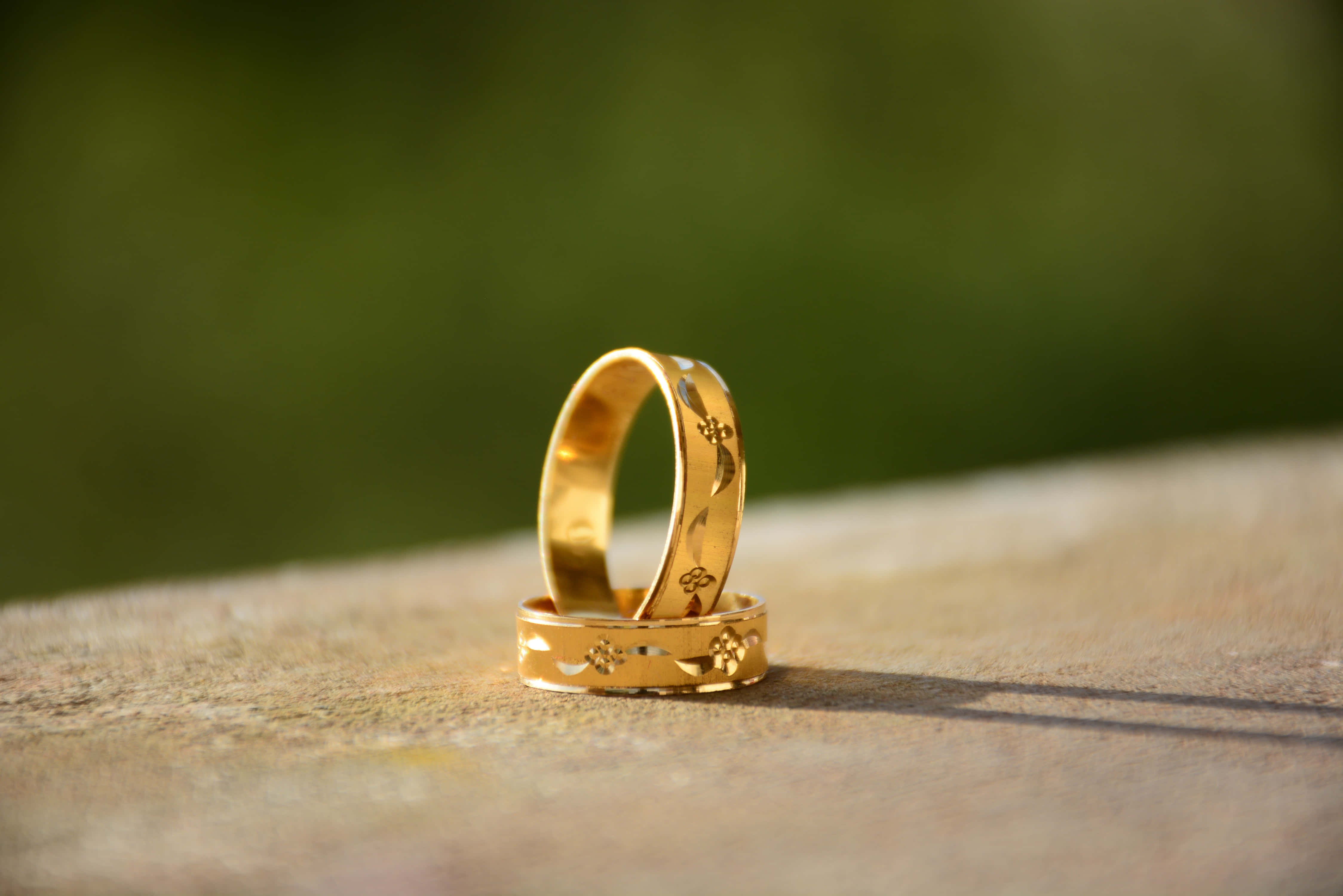 Wedding Rings 桌布 壁紙 — 兩枚帶有花卉雕刻的金色結婚戒指，在柔和的綠色背景下，放置在陽光照射的石頭表面上。