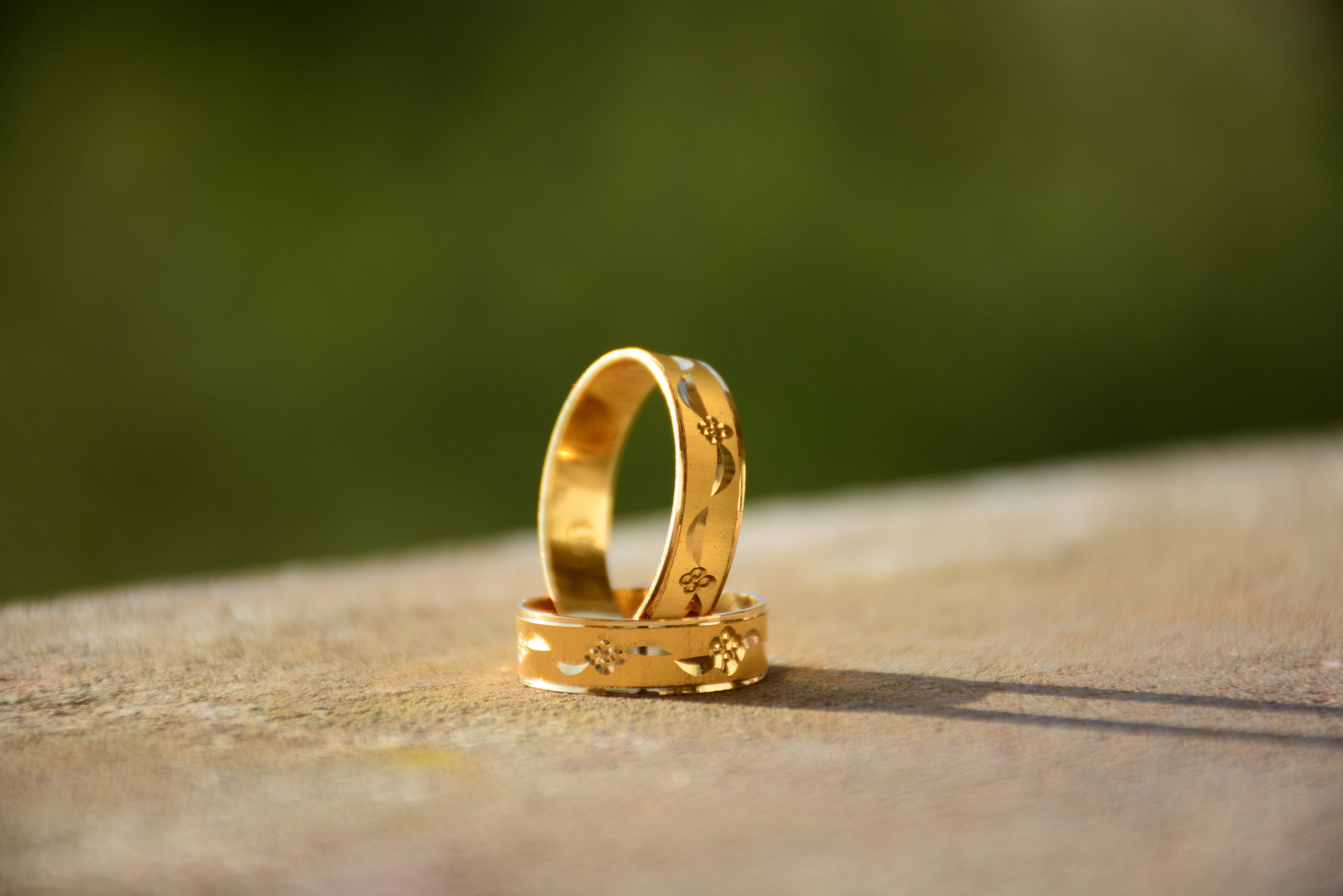 Gold Jewellery 桌布 壁紙 — 兩枚帶有花卉雕刻的金色結婚戒指，在柔和綠色背景前，放置在陽光照射的木質表面上。
