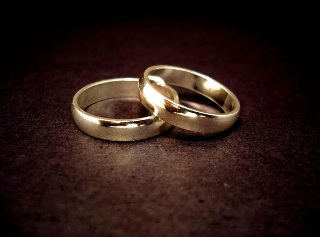 結婚戒指 桌布 壁紙 — 兩枚拋光金色結婚戒指在柔和的定向燈光下交纏在深色紋理表面上。