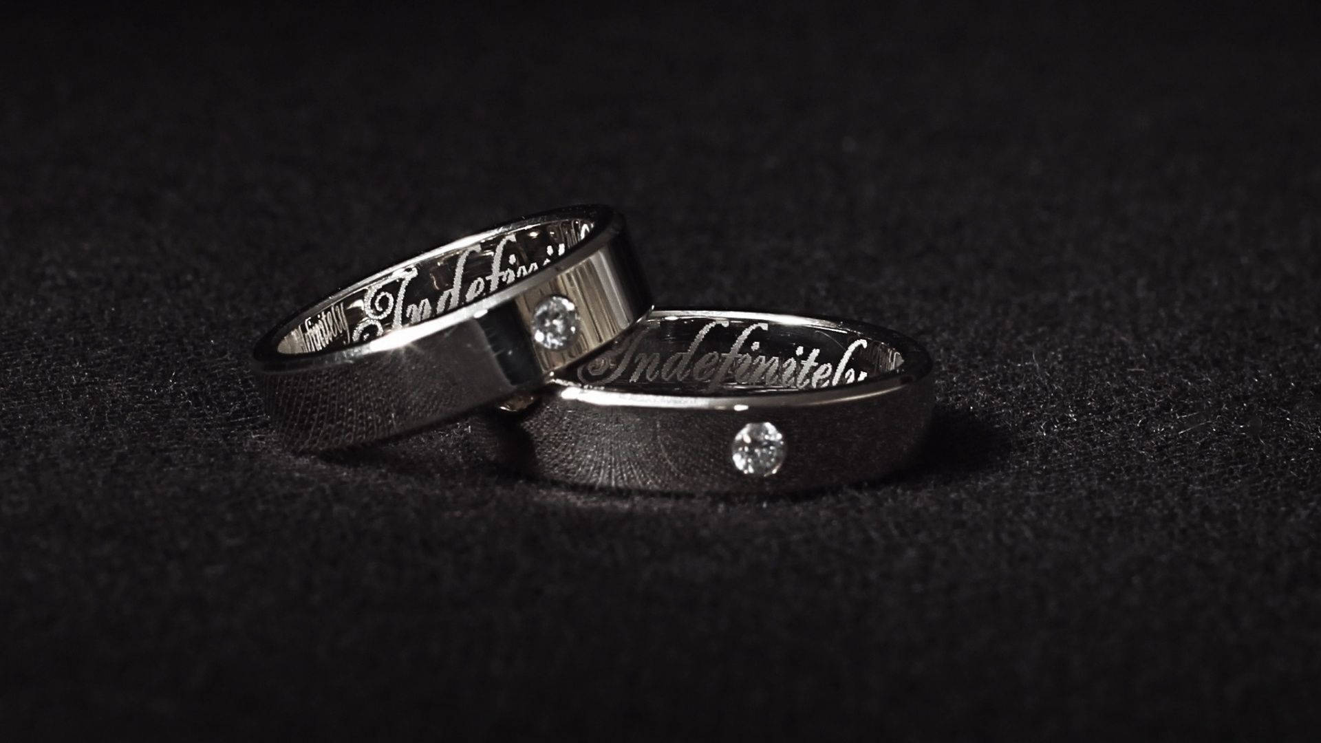 Wedding Rings 桌布 壁紙 — 兩枚時尚的銀色結婚戒指，鑲嵌鑽石並刻有「Indefinitely」，放置在柔軟的黑色紋理表面上。