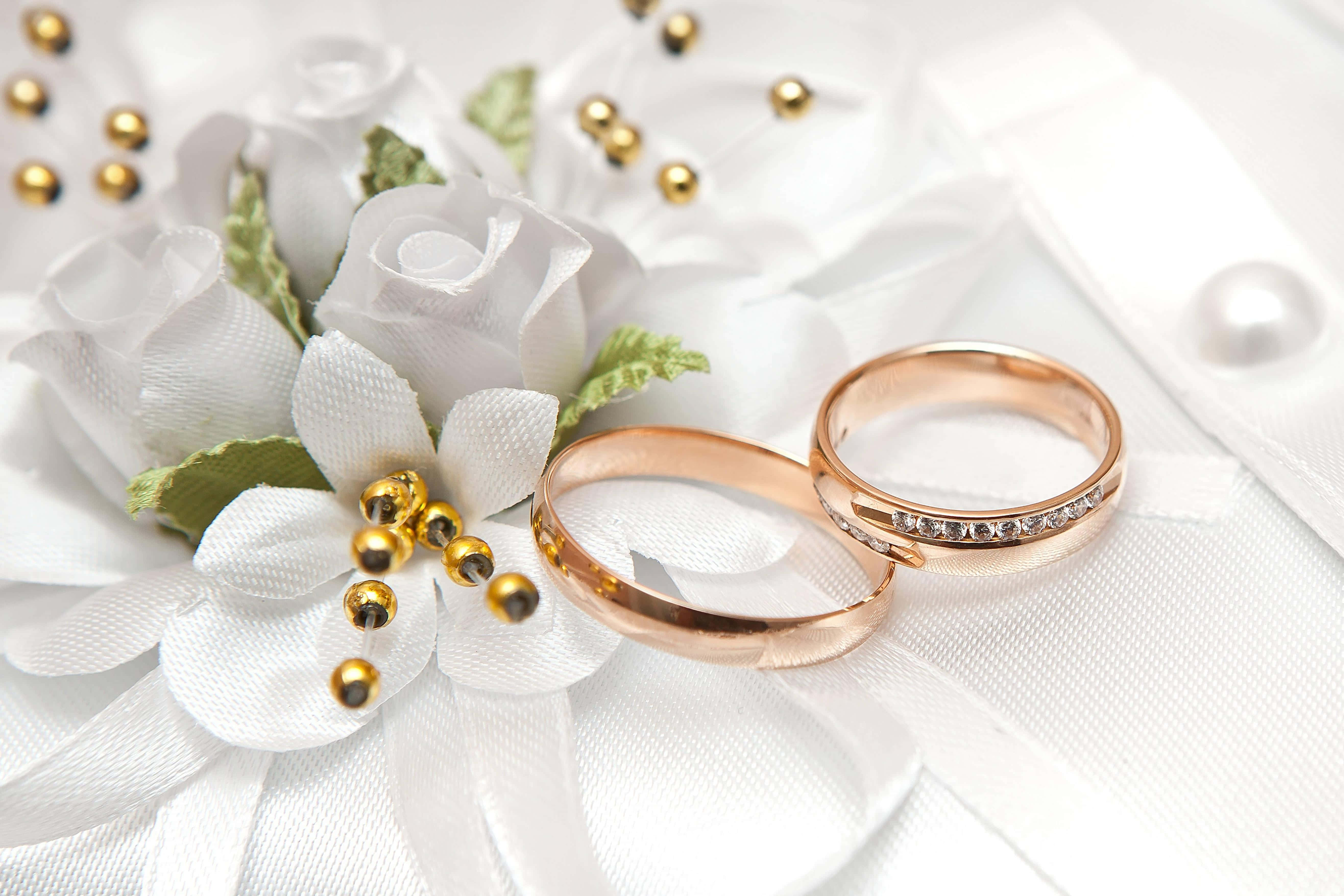 Wedding Rings 桌布 壁紙 — 兩枚玫瑰金結婚戒指放在白色布藝玫瑰和金珠旁邊，置於柔軟的白色緞面之上。