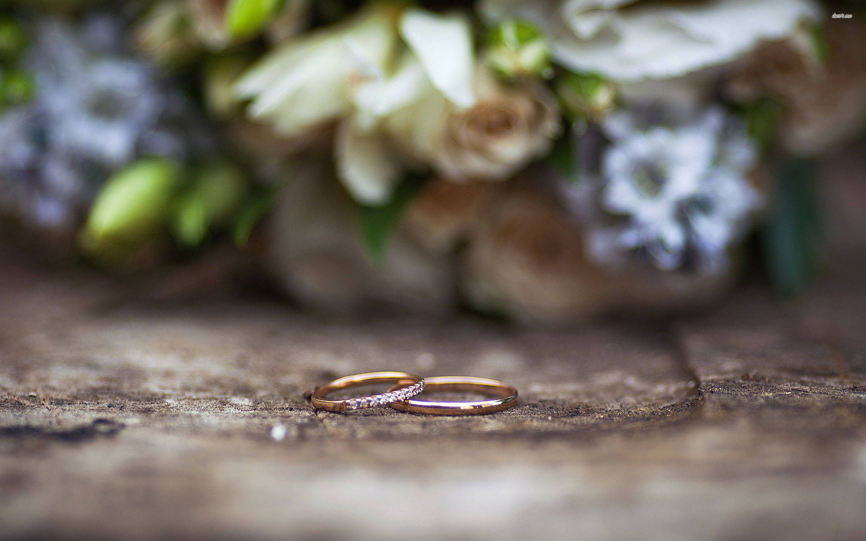 Wedding Rings 桌布 壁紙 — 兩枚玫瑰金結婚戒指放在風化木上，背景有柔和焦點的花束。