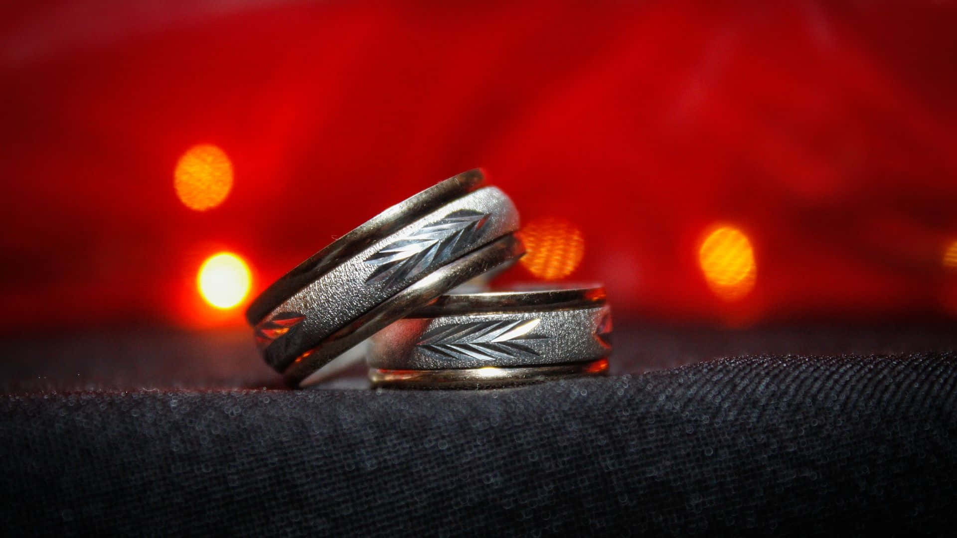Wedding Rings 桌布 壁紙 — 兩枚精雕細琢的銀色結婚戒指放在深色布料上，背景是鮮豔的紅色散景。