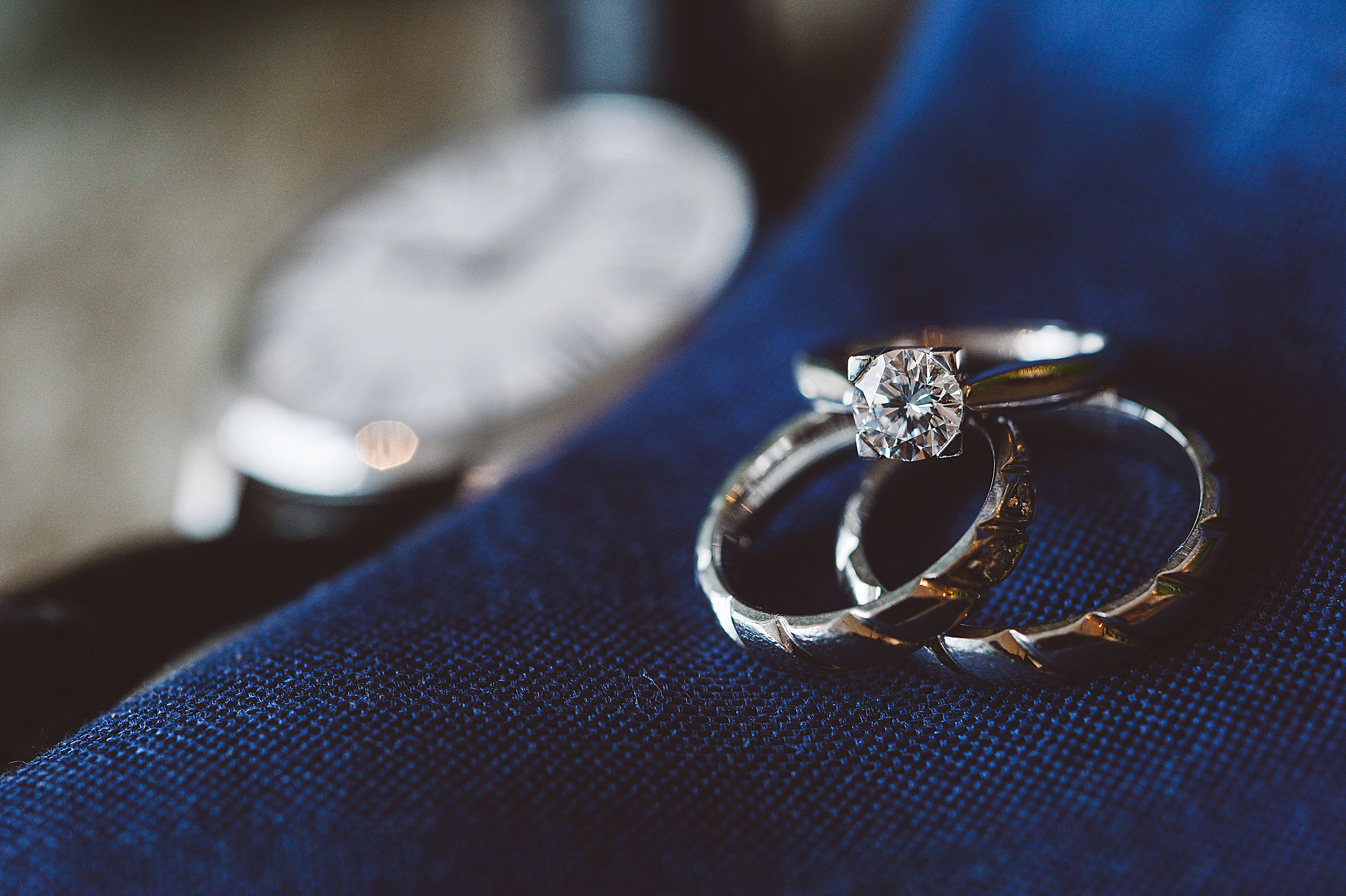 鑽石 桌布 壁紙 — 兩枚結婚戒指，一枚帶鑽石和紋理戒環，放在深藍色布料上，旁邊有一隻模糊的手錶。