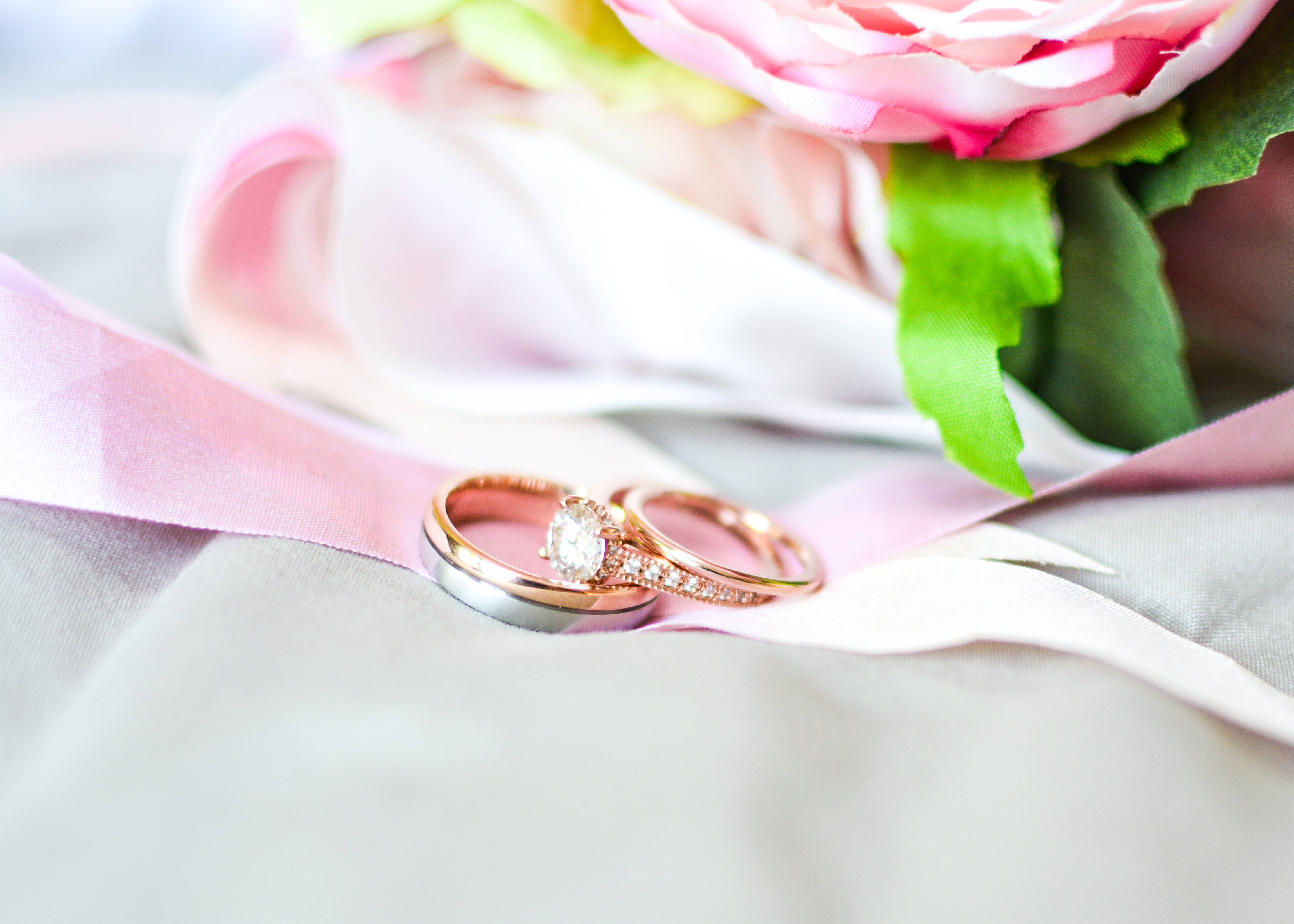 玫瑰金 桌布 壁紙 — 兩枚結婚戒指，一枚玫瑰金鑽石單石戒指和一枚素圈，放在柔軟的粉紅色絲帶上，旁邊是模糊的粉紅色牡丹花束。