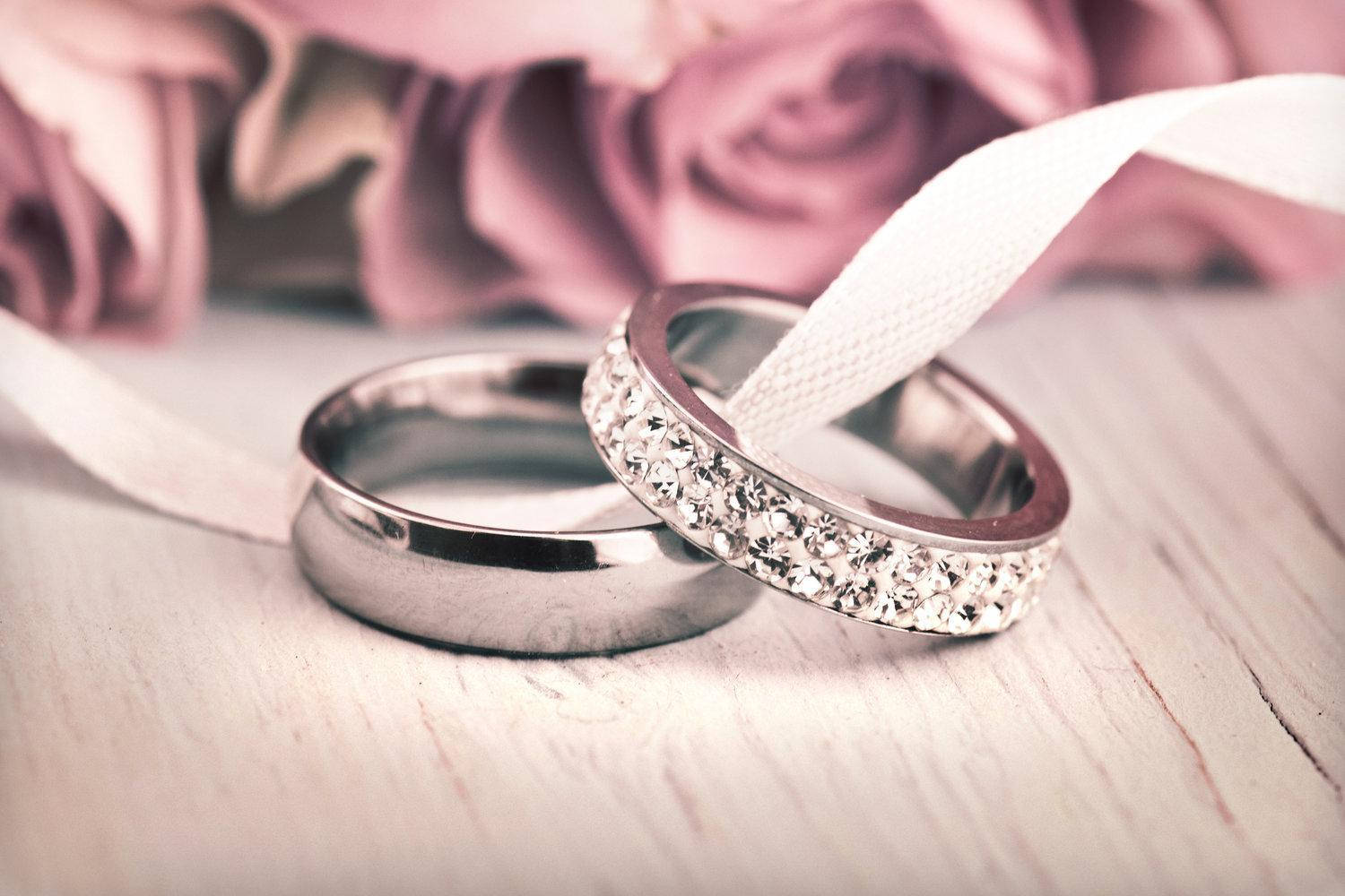 Wedding Rings 桌布 壁紙 — 兩枚結婚戒指，一枚素面，一枚鑲鑽，放在白色絲帶上，旁邊是柔和的粉紅玫瑰，置於木質表面。