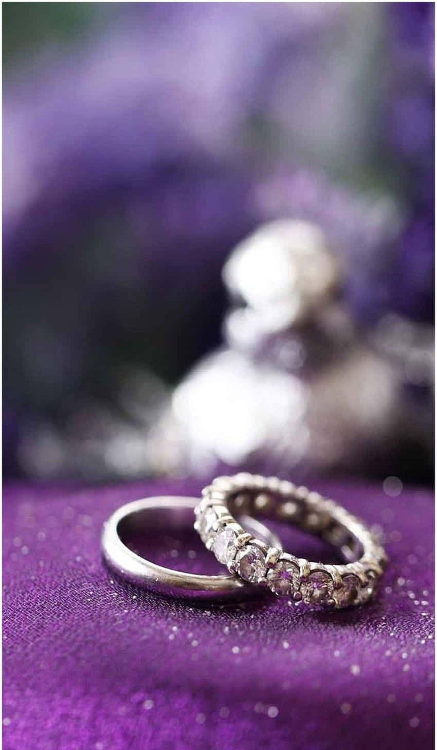 Marriage 桌布 壁紙 — 兩枚結婚戒指，一枚素面，一枚鑲鑽，放在閃爍的紫色布料上，背景是模糊的花卉。